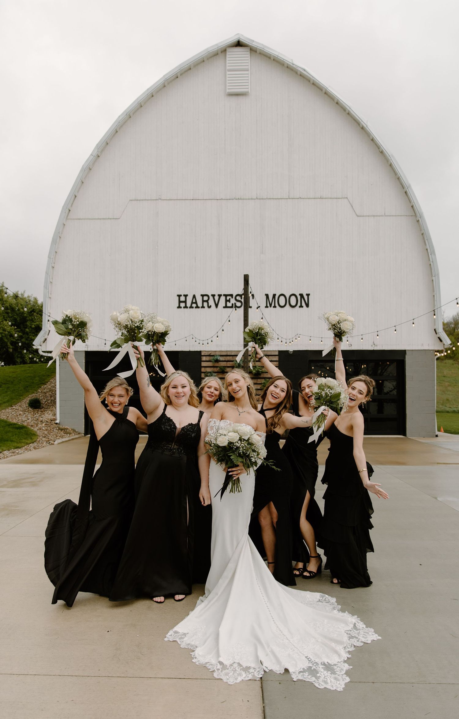 Bride in white gown with bridesmaids in black dresses celebrating outside Harvest Moon barn venue.