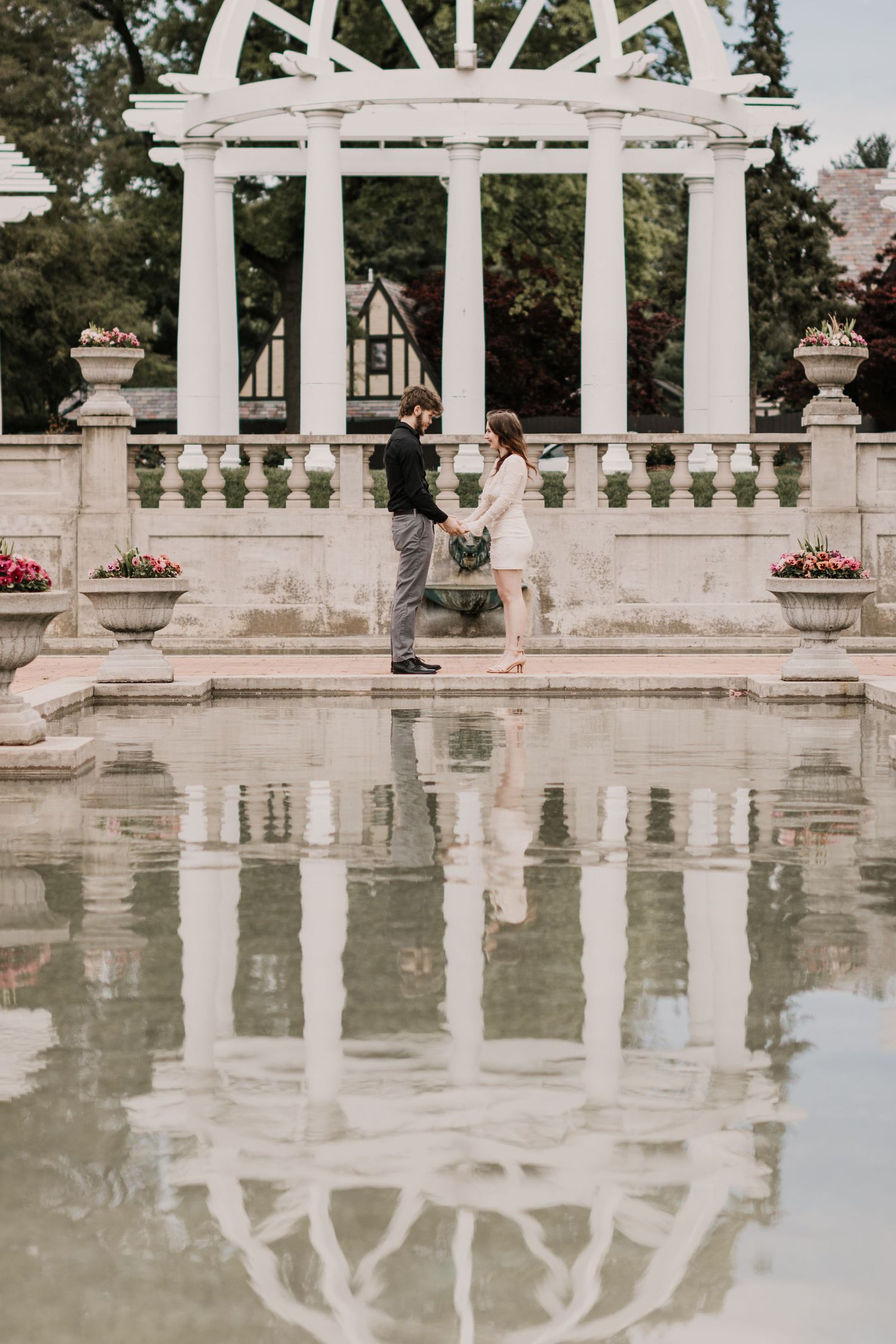 A couple stands under a white pergola with ornate columns reflected in a pool of water.