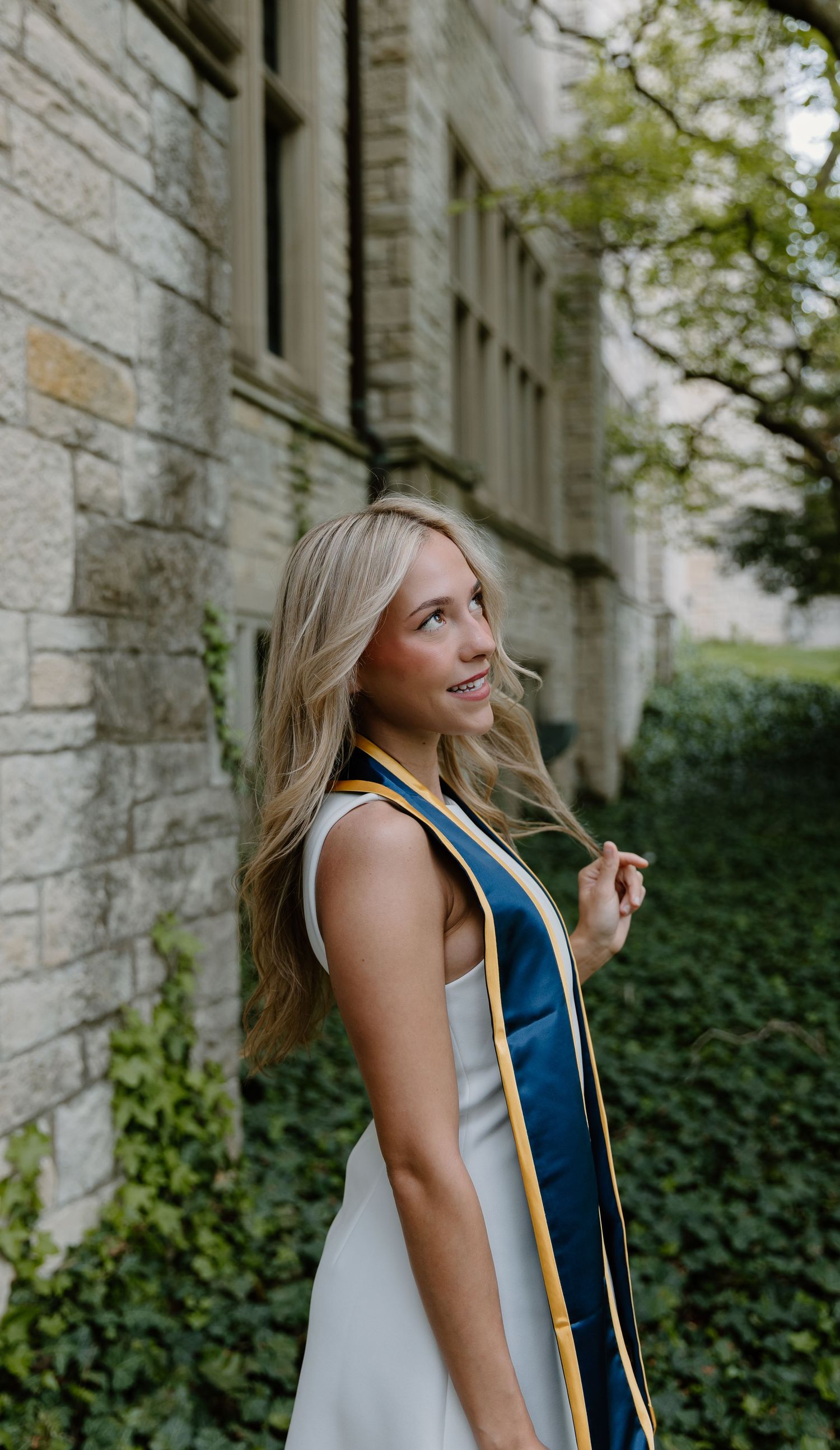 Smiling blonde graduate wearing a navy and gold sash poses outdoors beside a stone building with ivy.