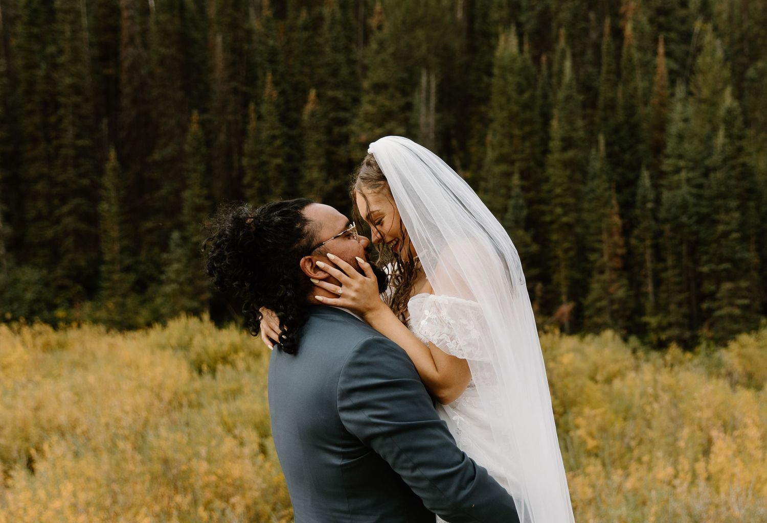 A romantic wedding couple shares a kiss in front of a scenic mountain landscape with evergreen trees and golden grass.