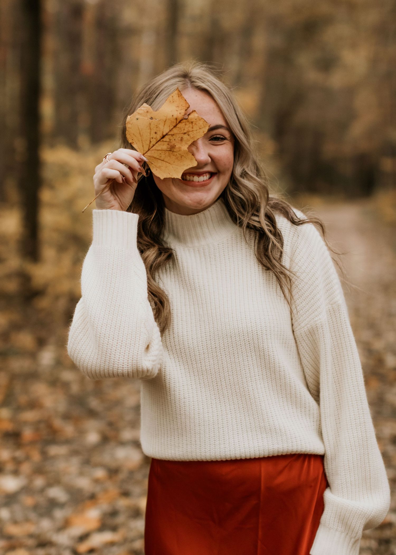 Playful autumn portrait with person holding a fall leaf wearing cream sweater and rust skirt.