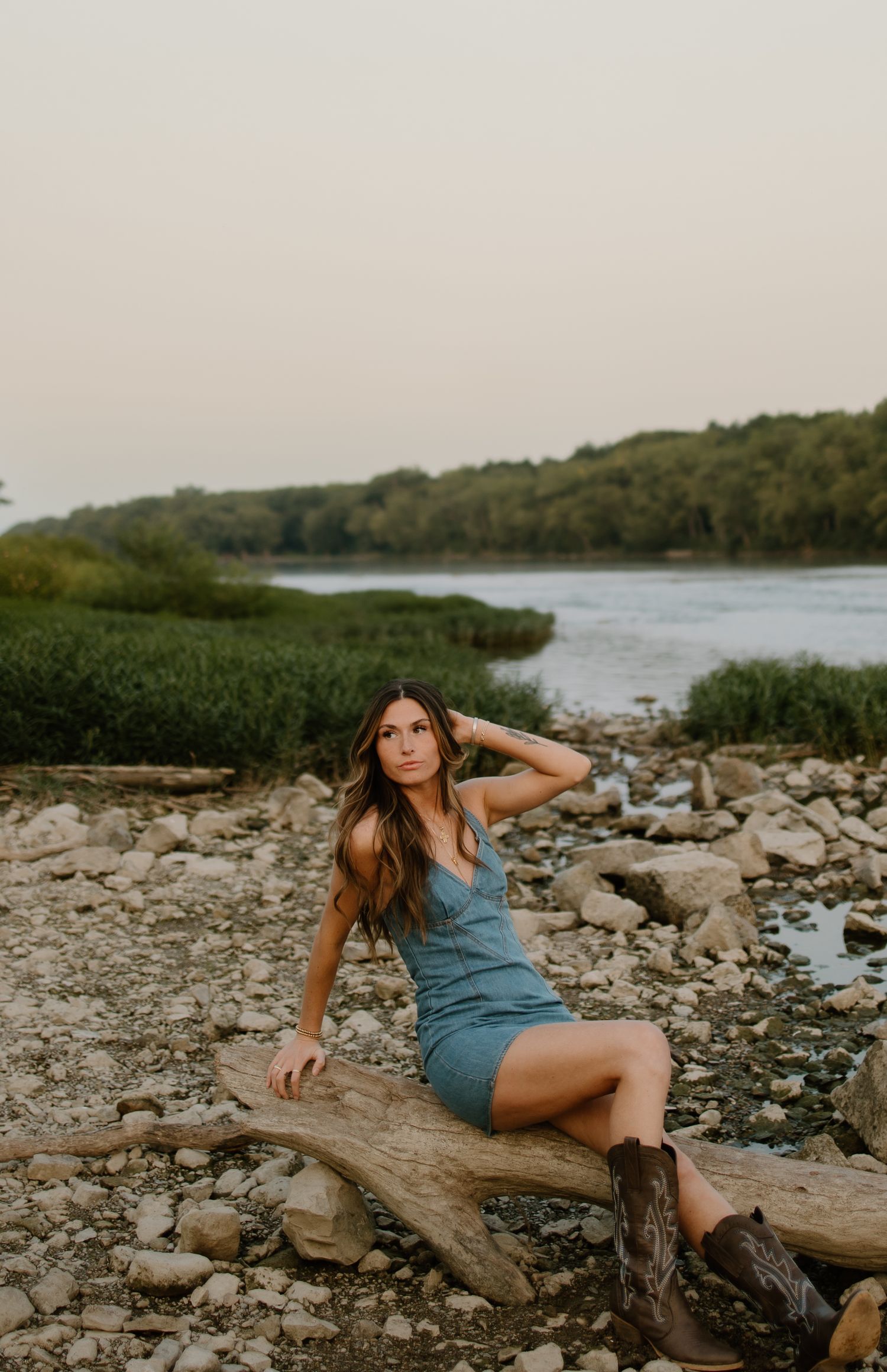 Woman in blue dress and cowboy boots sitting on driftwood by rocky riverbank at sunset.
