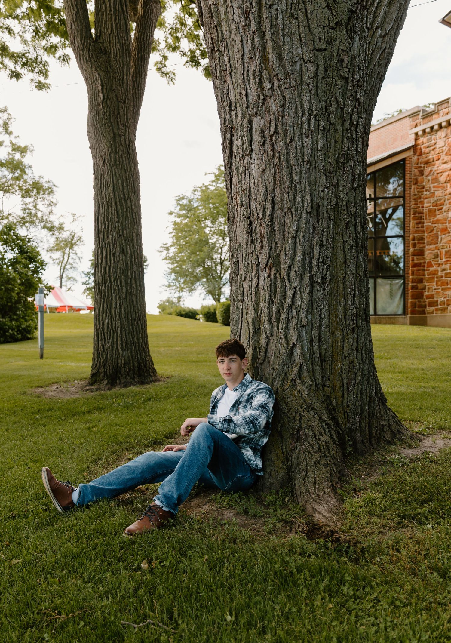 Young man in plaid shirt and jeans sitting against large tree trunk on green lawn near brick building.