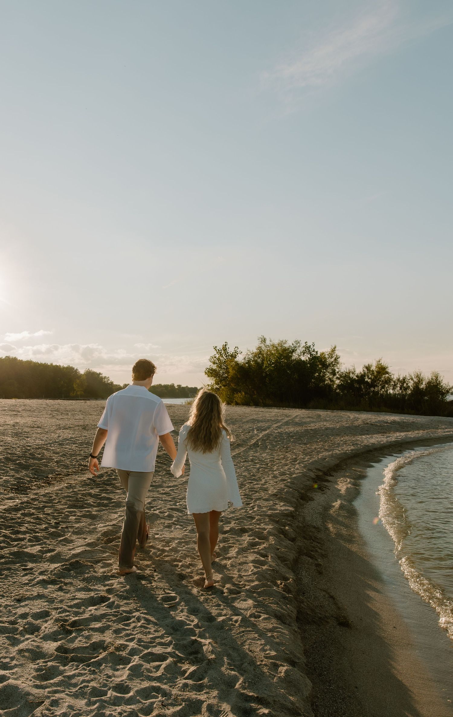A couple walks hand-in-hand along a sandy beach at sunset, wearing white outfits with trees in the background.