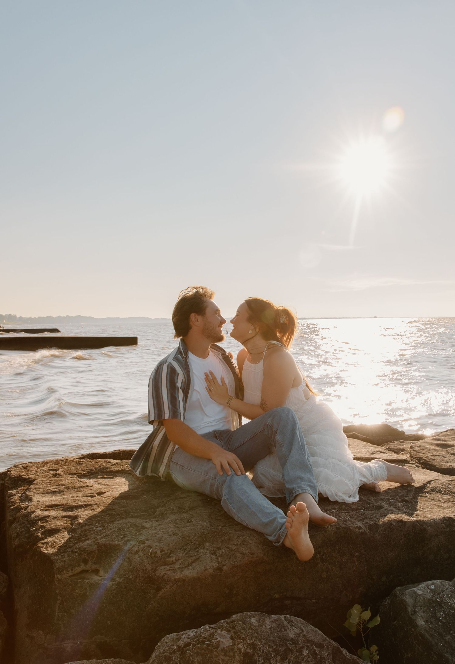 Couple sitting on rocks by the ocean, leaning in for a kiss with golden sunlight glowing behind them.