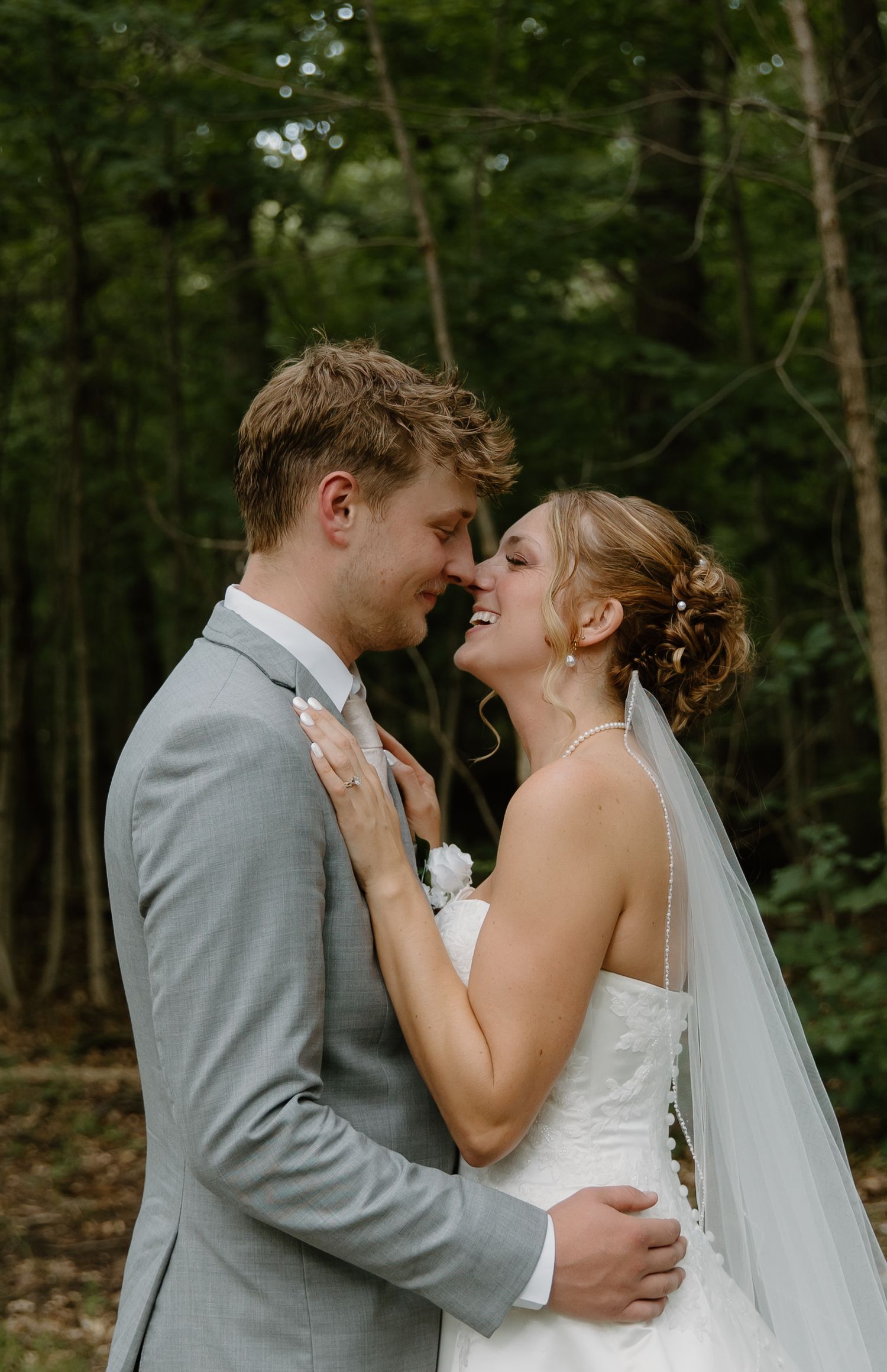 A smiling bride and groom share a romantic moment in a lush forest setting, with the bride in a white gown and veil.
