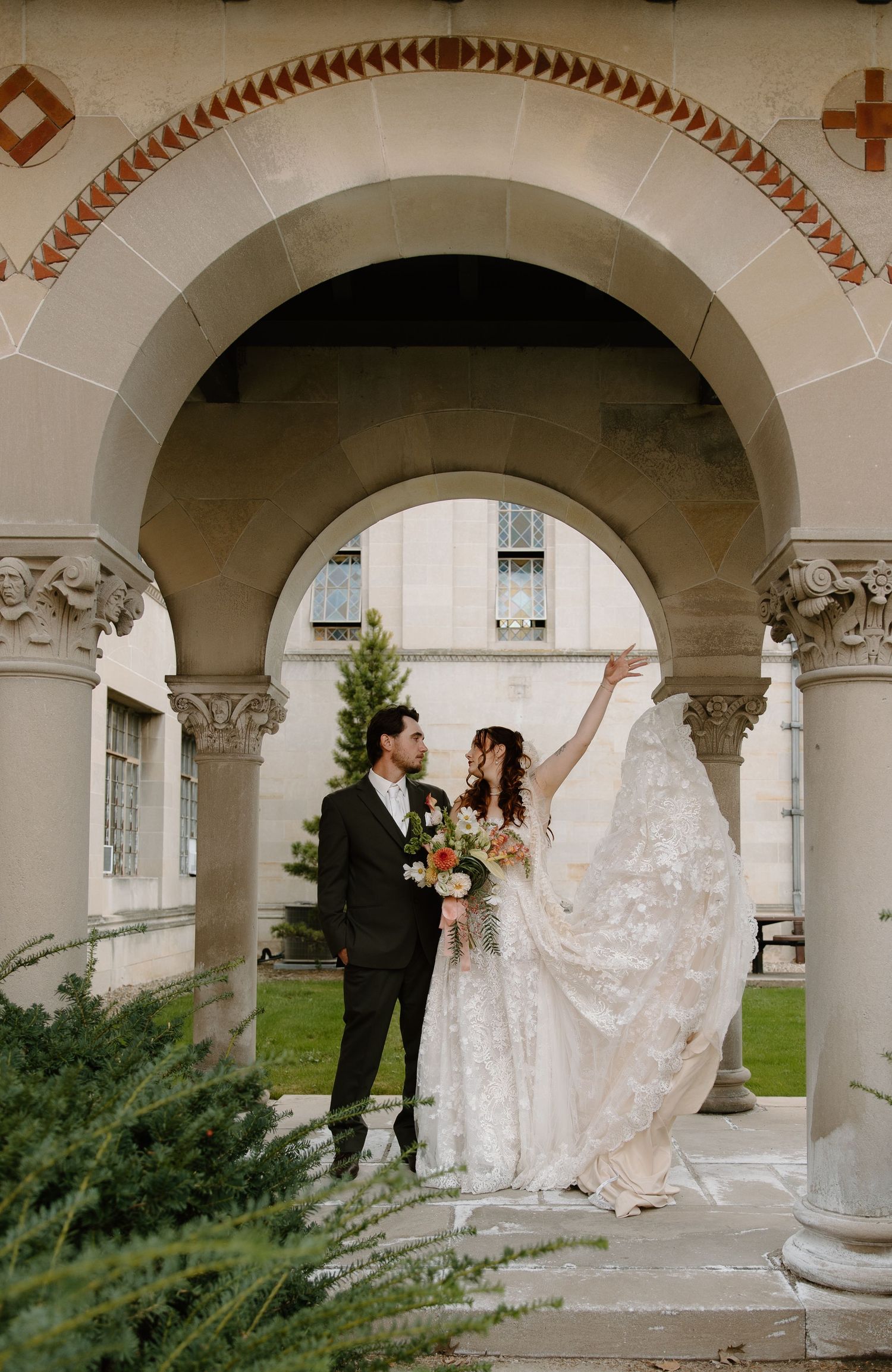 Bride and groom stand under stone arch at elegant venue, bride's veil flowing as she reaches upward, holding floral bouquet.