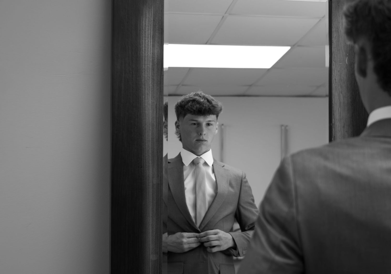 A person in a suit adjusts their tie while looking in a mirror in a hallway.