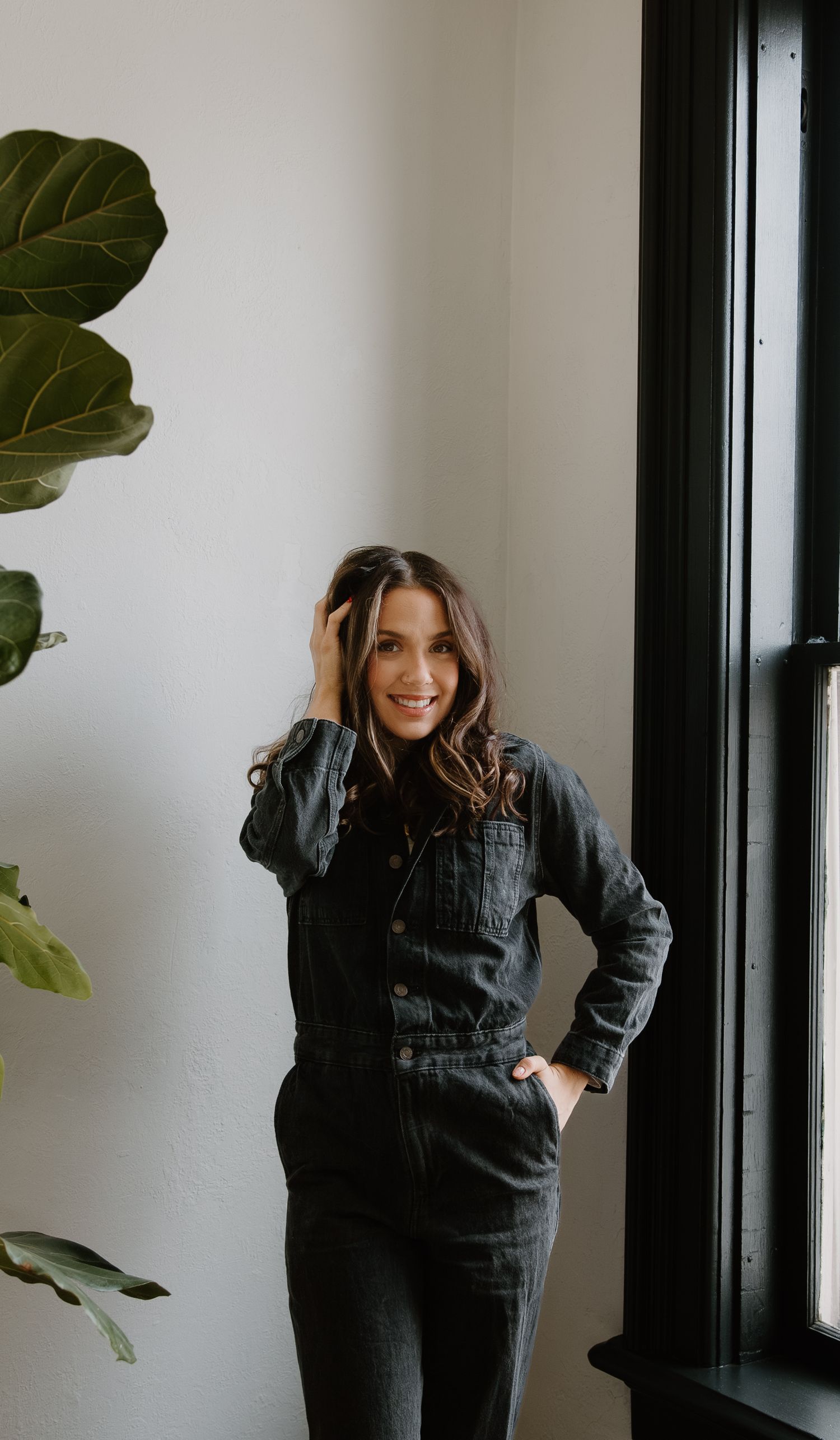 Smiling woman in dark denim jumpsuit poses near window with fiddle leaf fig plant in bright modern interior.