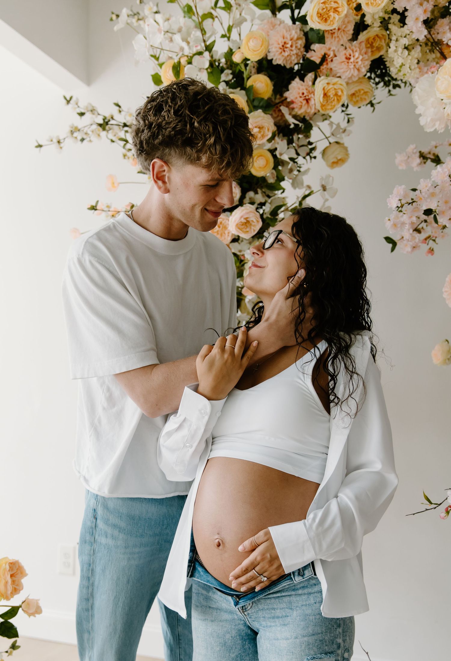 Couple sharing a tender moment during a maternity photoshoot with floral backdrop, woman cradling baby bump.