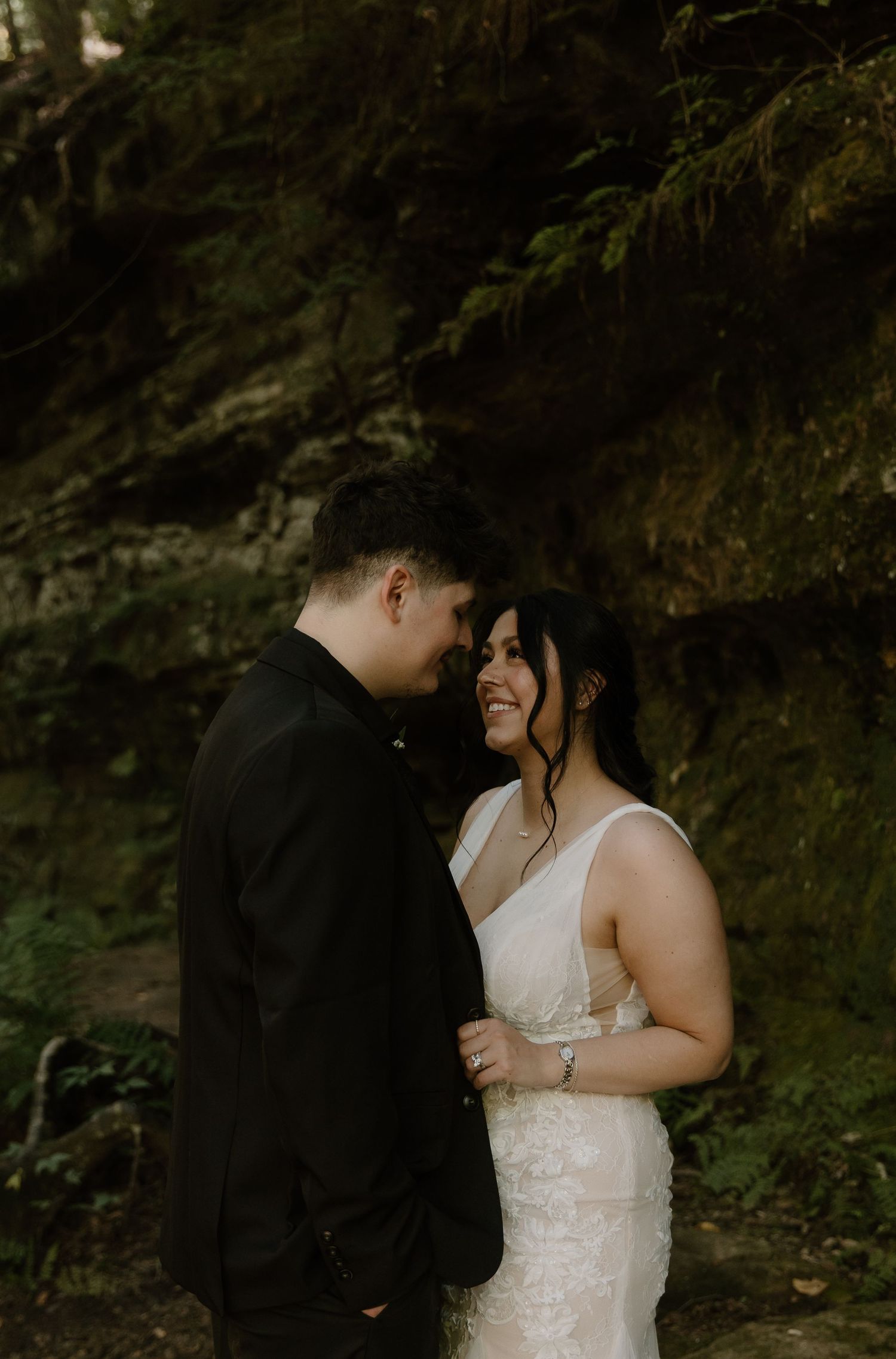 A couple in wedding attire shares a romantic moment in a lush forest, the bride smiling joyfully in a white lace gown.