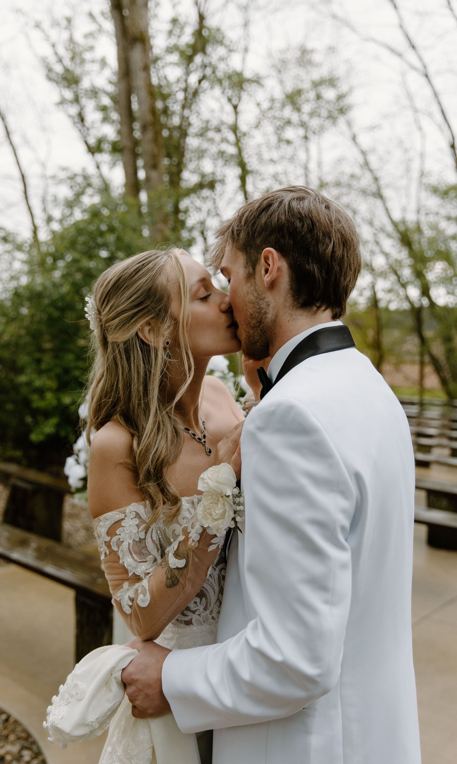 Bride and groom sharing a romantic kiss outdoors, bride in lace off-shoulder gown, groom in white tuxedo jacket.
