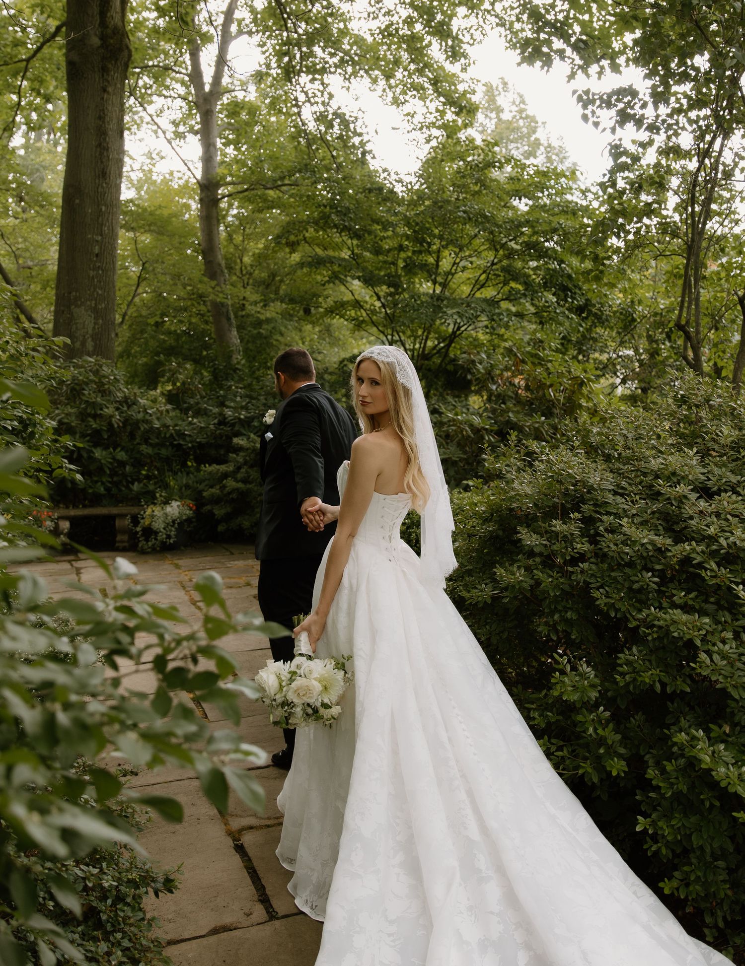 Bride in a strapless lace gown holds bouquet on a garden path, glancing back while groom leads the way.