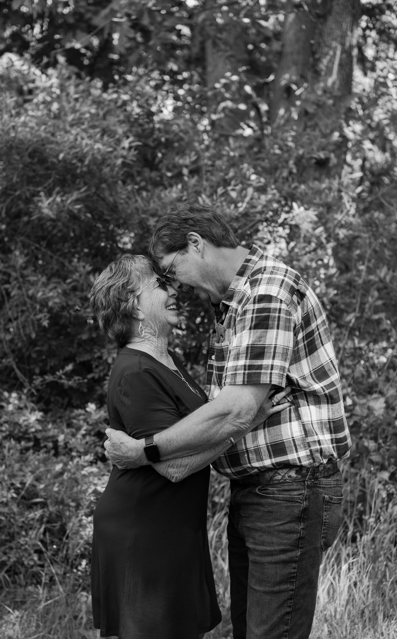 A tender black and white portrait of a couple embracing outdoors in a natural wooded setting.