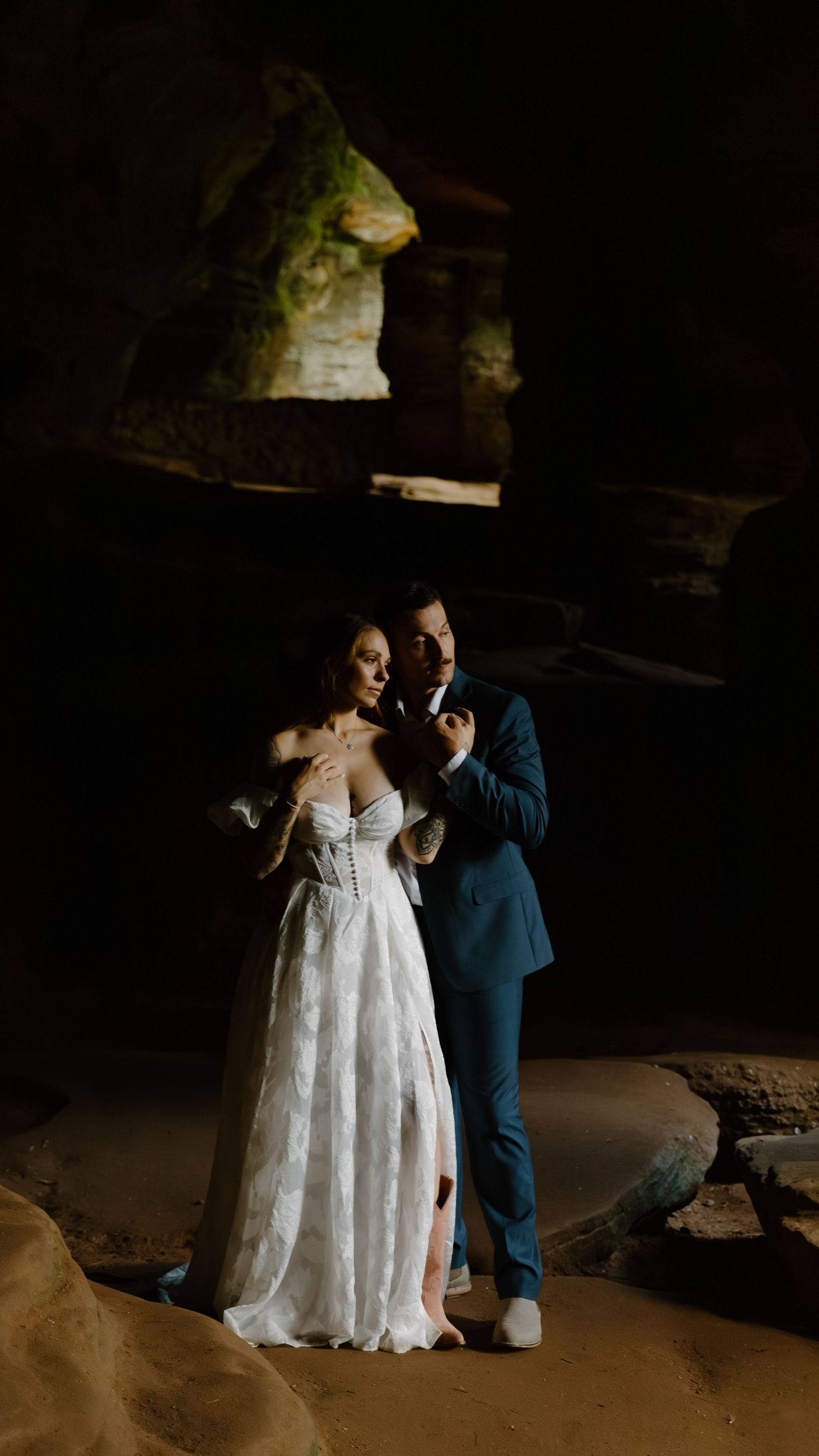A couple in wedding attire stands together in a dramatic cave setting with moody lighting and natural rock formations.