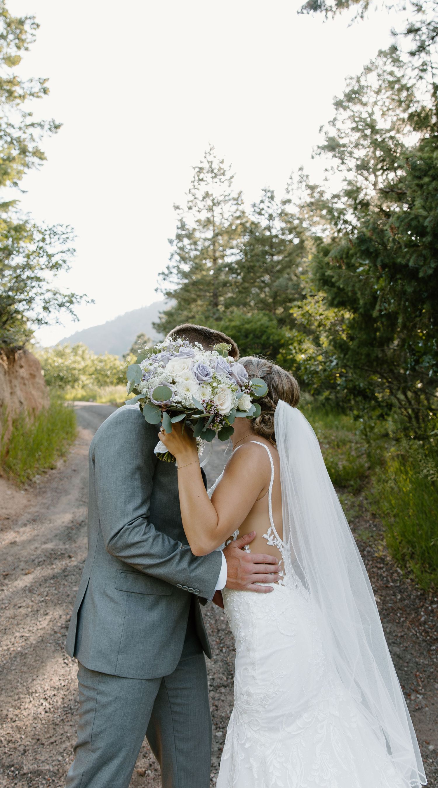 Newlyweds share an intimate kiss on a mountain trail with a bouquet of white and green flowers.