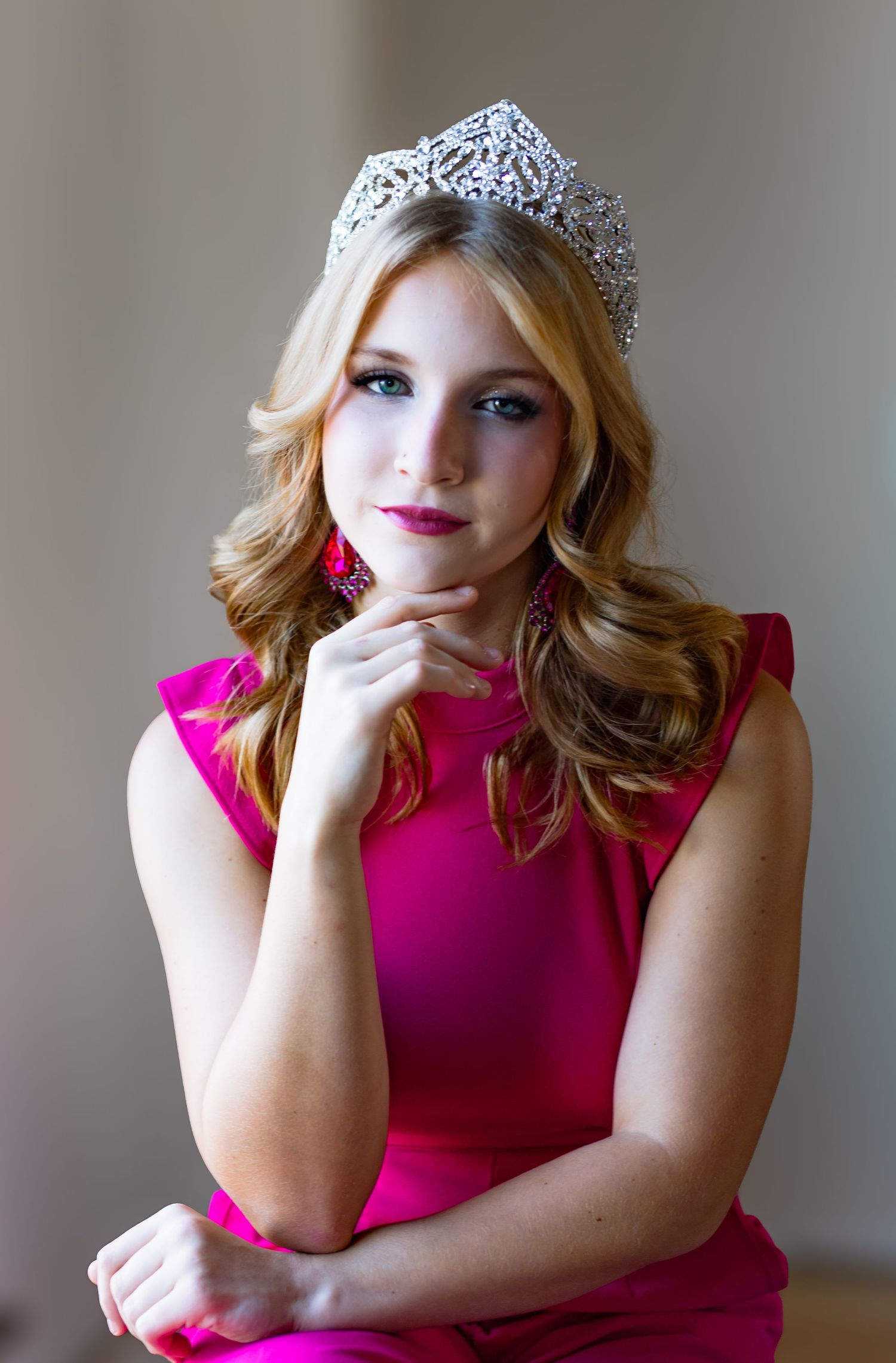 Glamorous portrait with sparkling crown and fuchsia dress against neutral backdrop.