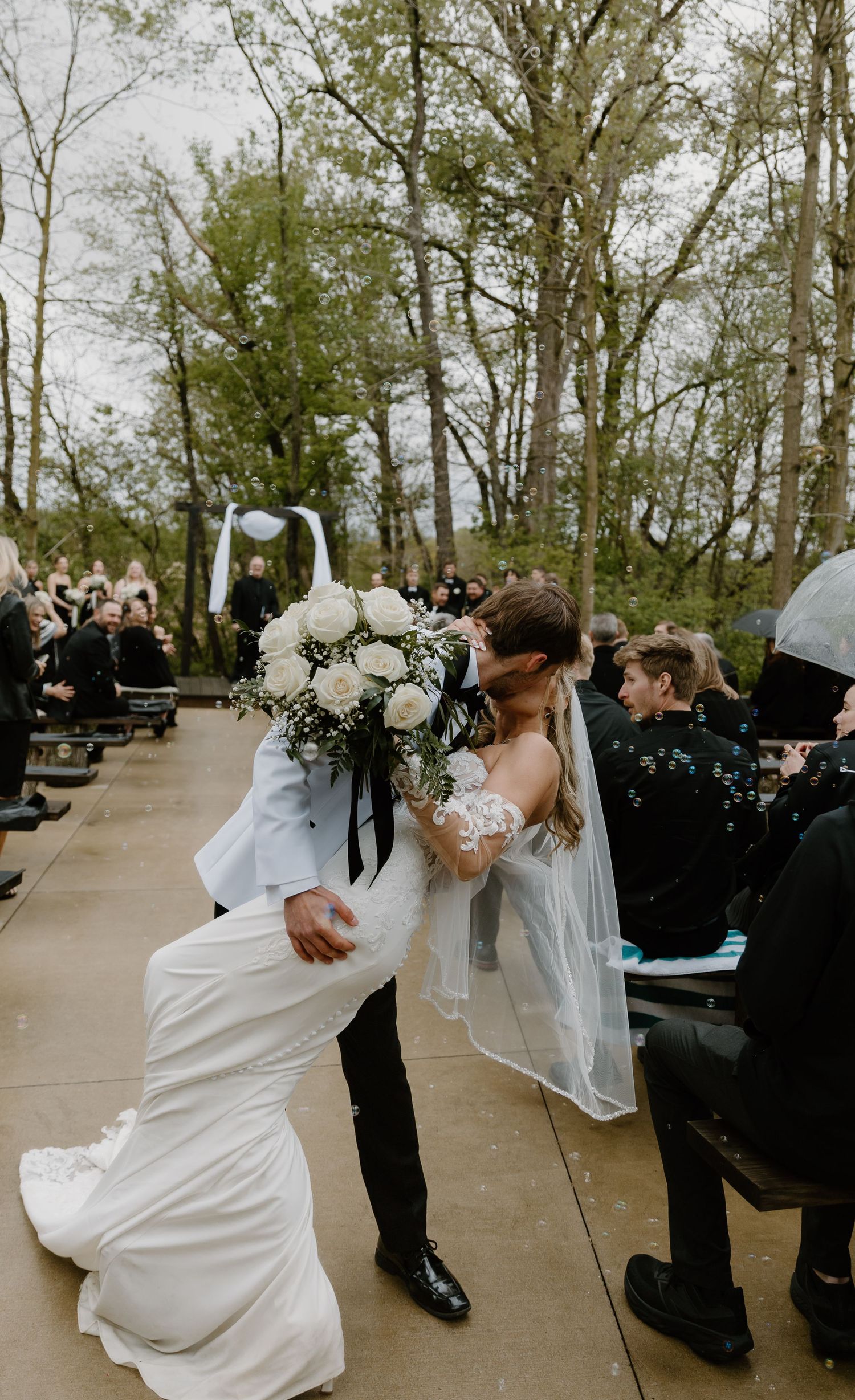 Bride and groom share romantic dip kiss while walking down outdoor wedding aisle surrounded by guests.