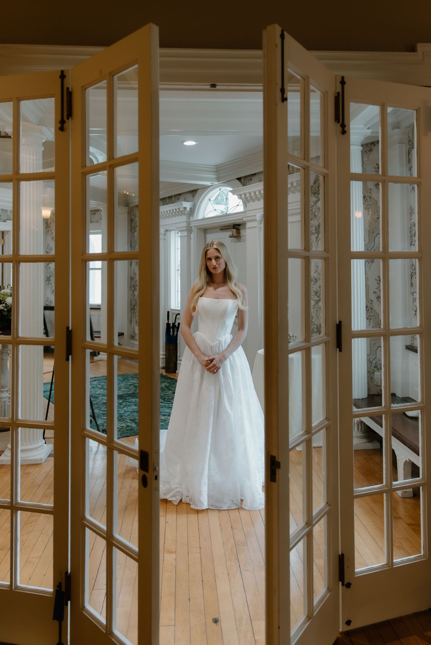Elegant bride in white strapless ballgown stands framed by French doors in classic mansion interior.