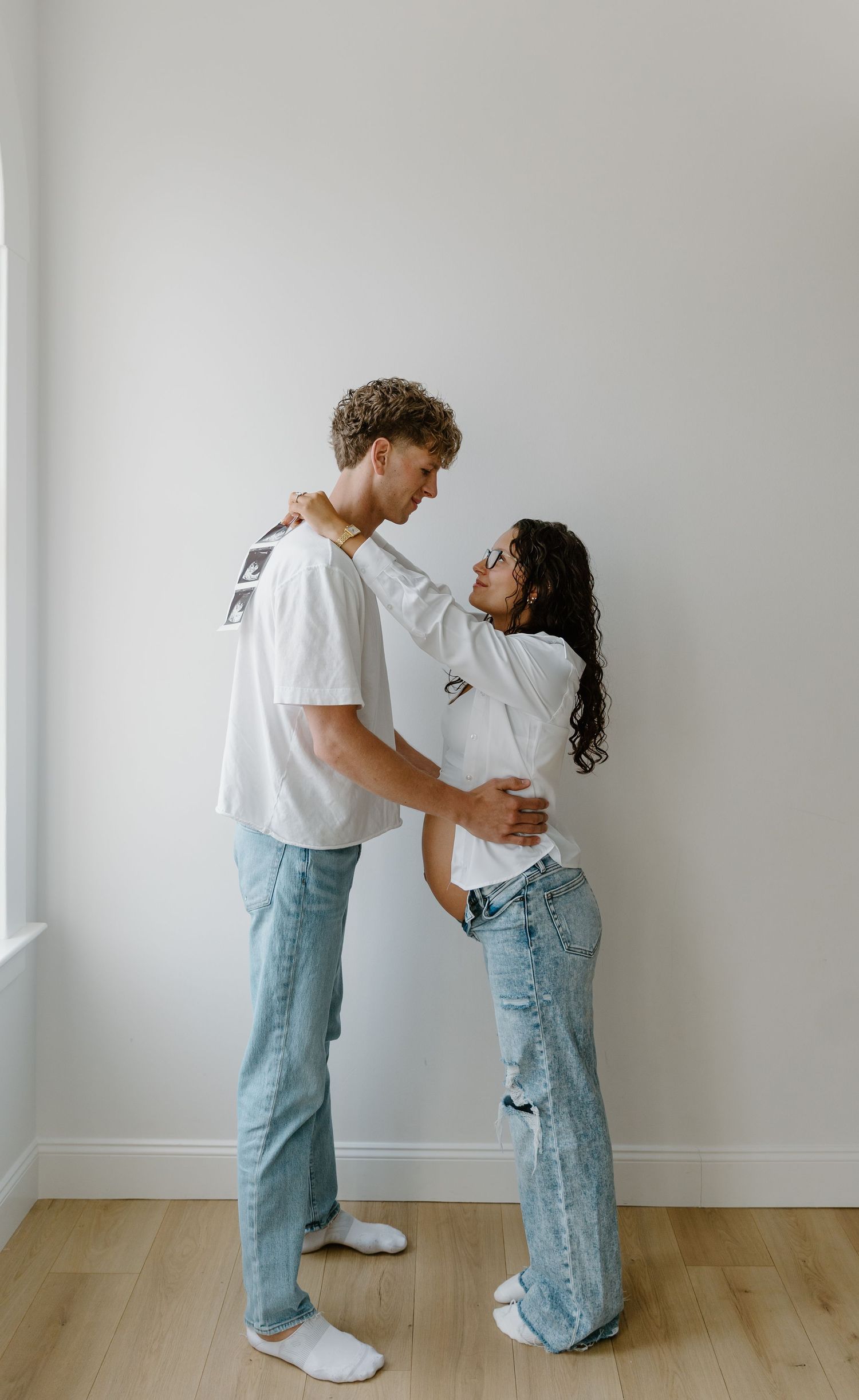 Expecting couple in matching white tees and jeans sharing a tender moment against a white wall.