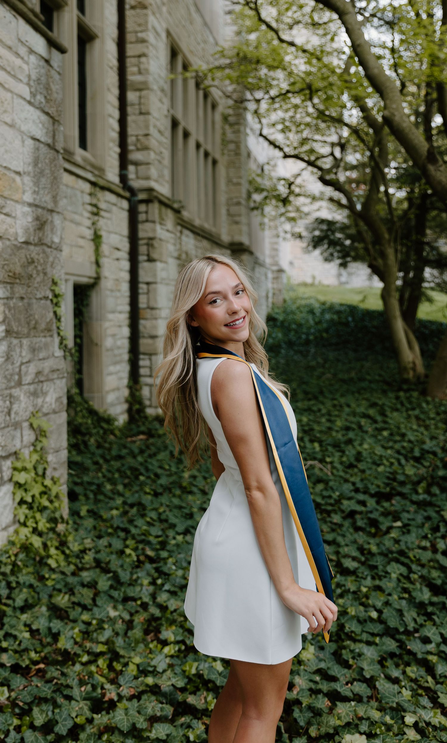 Smiling blonde woman in white dress with graduation sash poses outdoors near ivy-covered stone building.