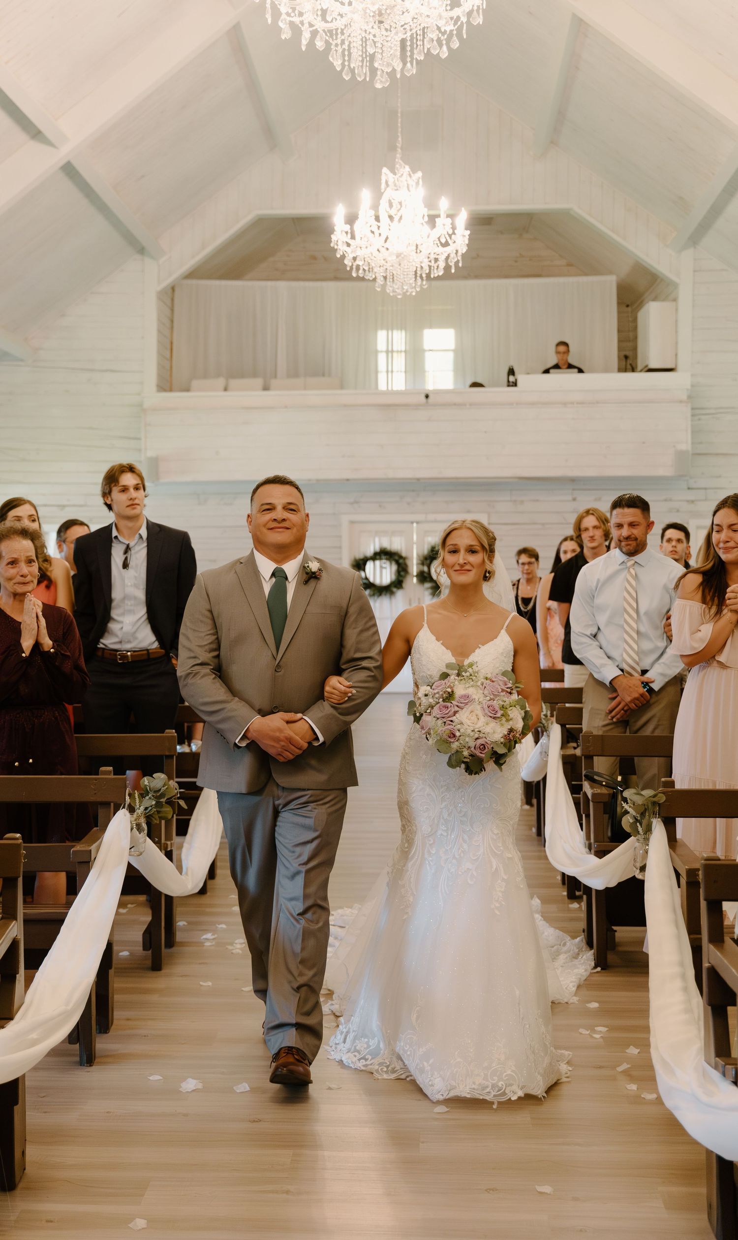 Wedding ceremony procession in a bright white chapel with elegant chandeliers and wooden flooring.