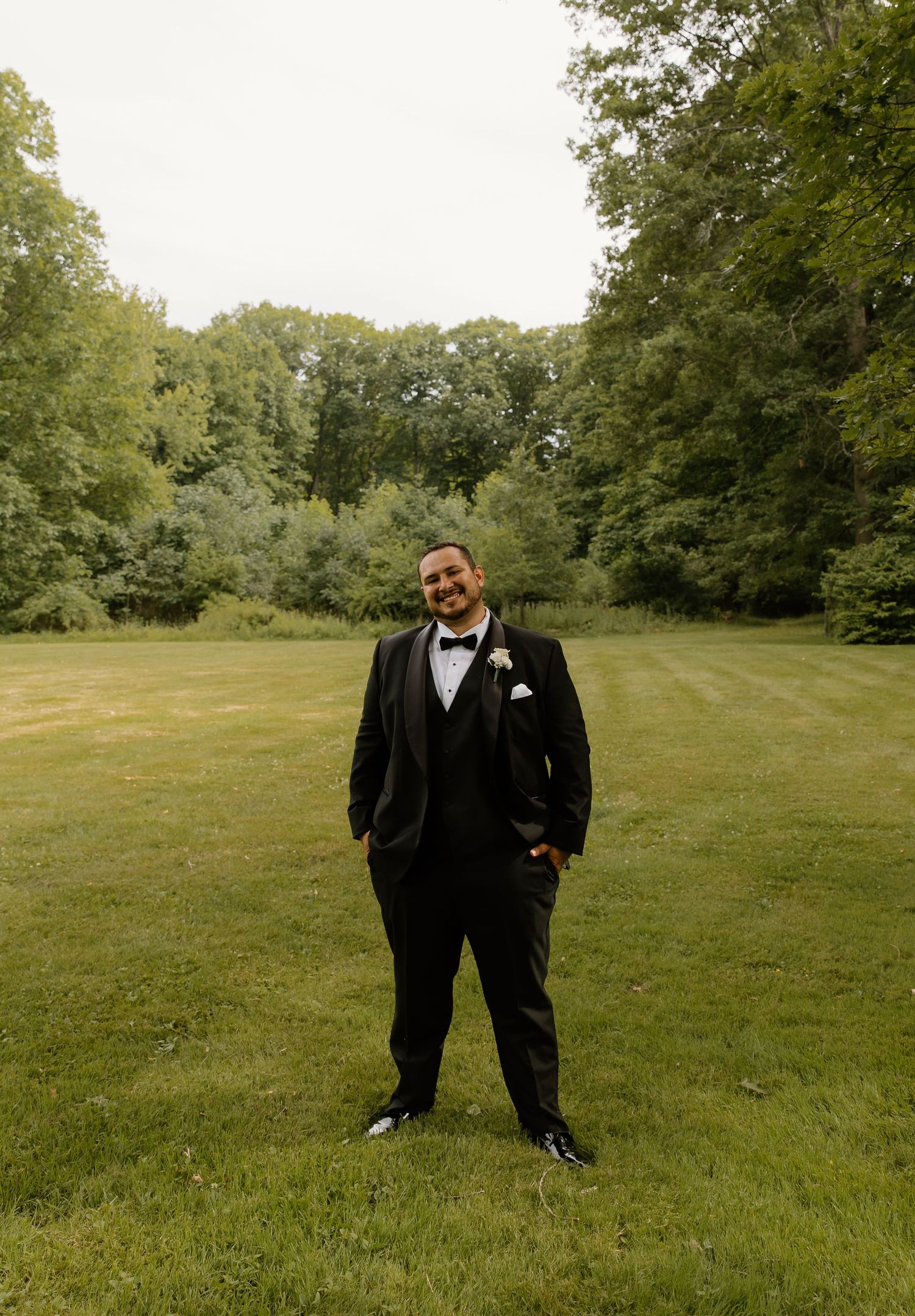 Smiling groom in a black tuxedo with bow tie standing on a lush green lawn before his wedding.