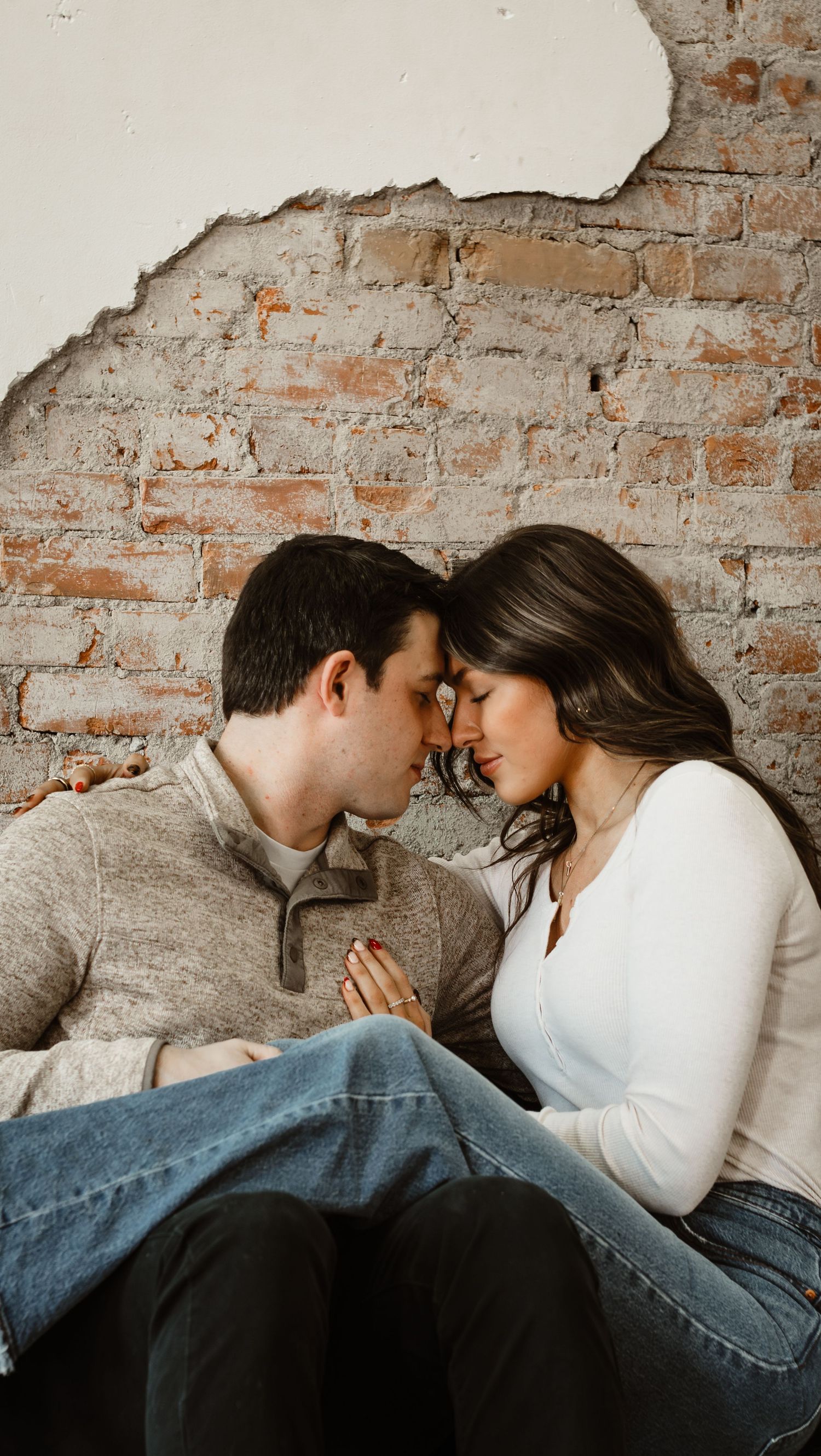 A couple shares an intimate moment on a couch against an exposed brick wall background.