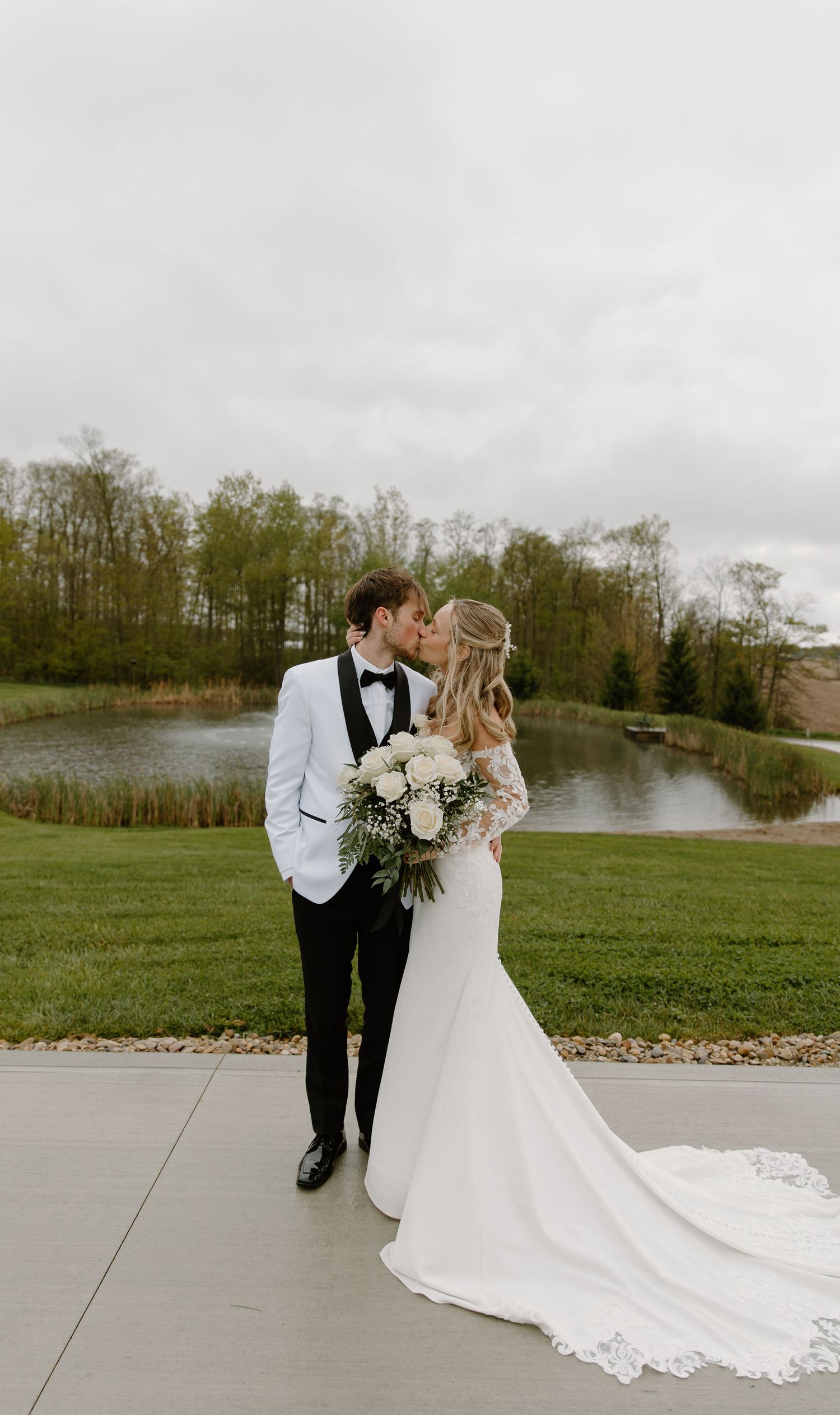 Newlyweds share a tender kiss in wedding attire, bride holding white bouquet near serene pond venue.