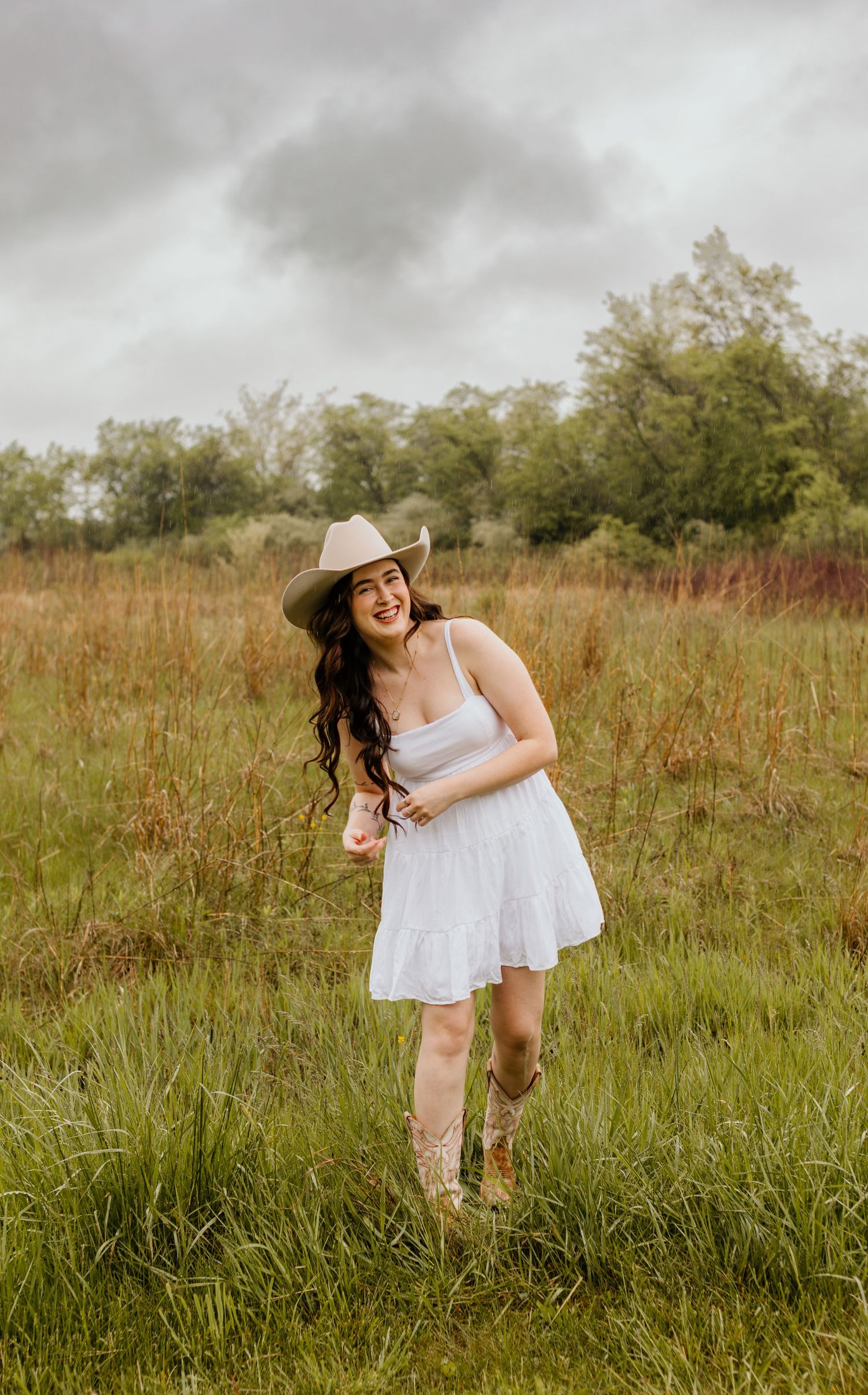 A person in a white sundress and cowboy hat dances joyfully in a pastoral field on a cloudy summer day.