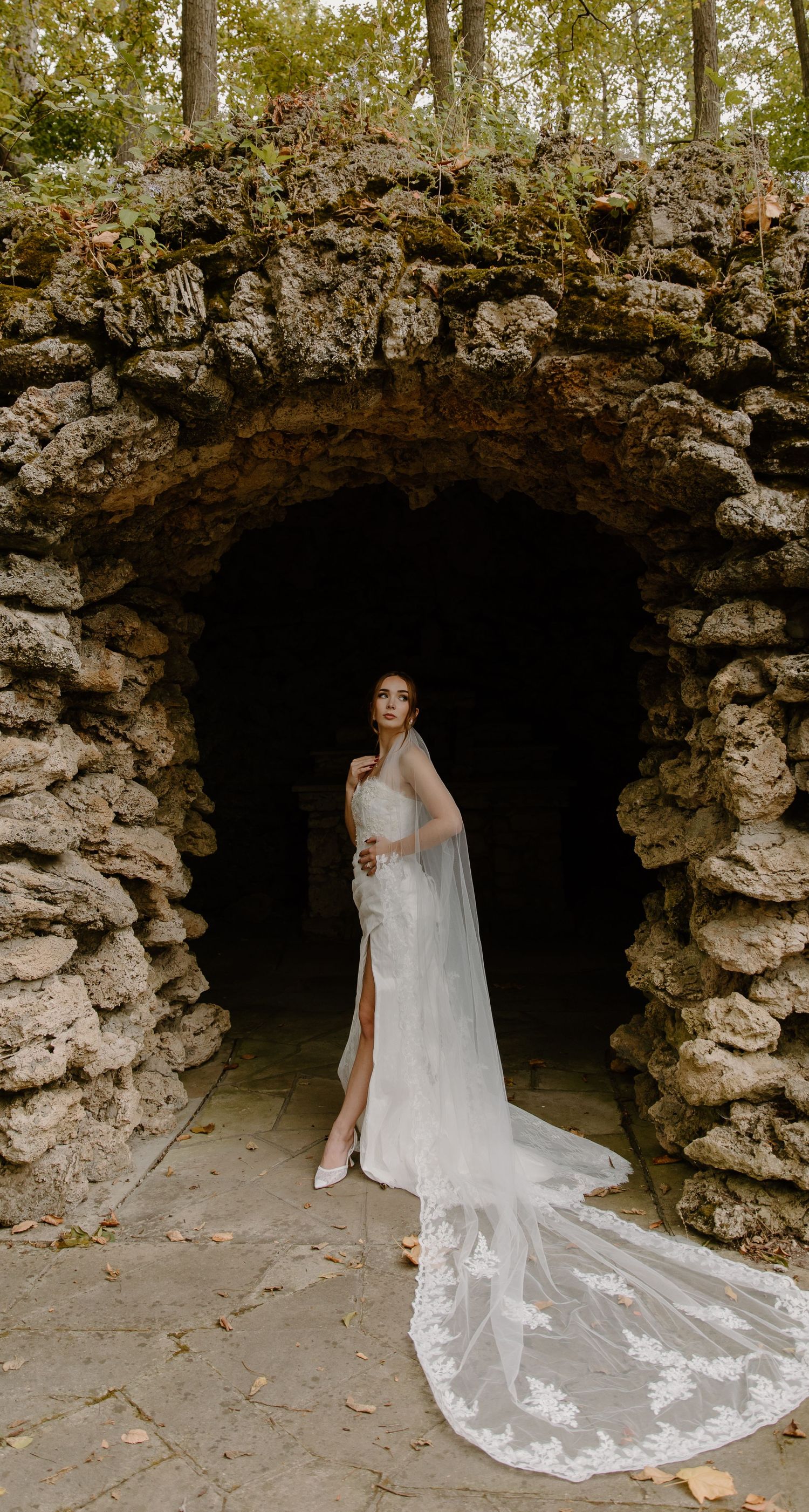 A bride in a flowing white wedding gown with a long veil poses elegantly in a stone grotto.