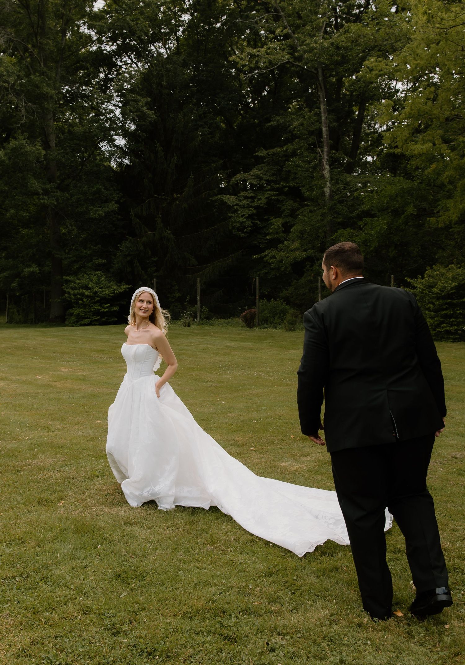 Bride in white gown with train stands on green lawn smiling at groom in black suit near forest backdrop.