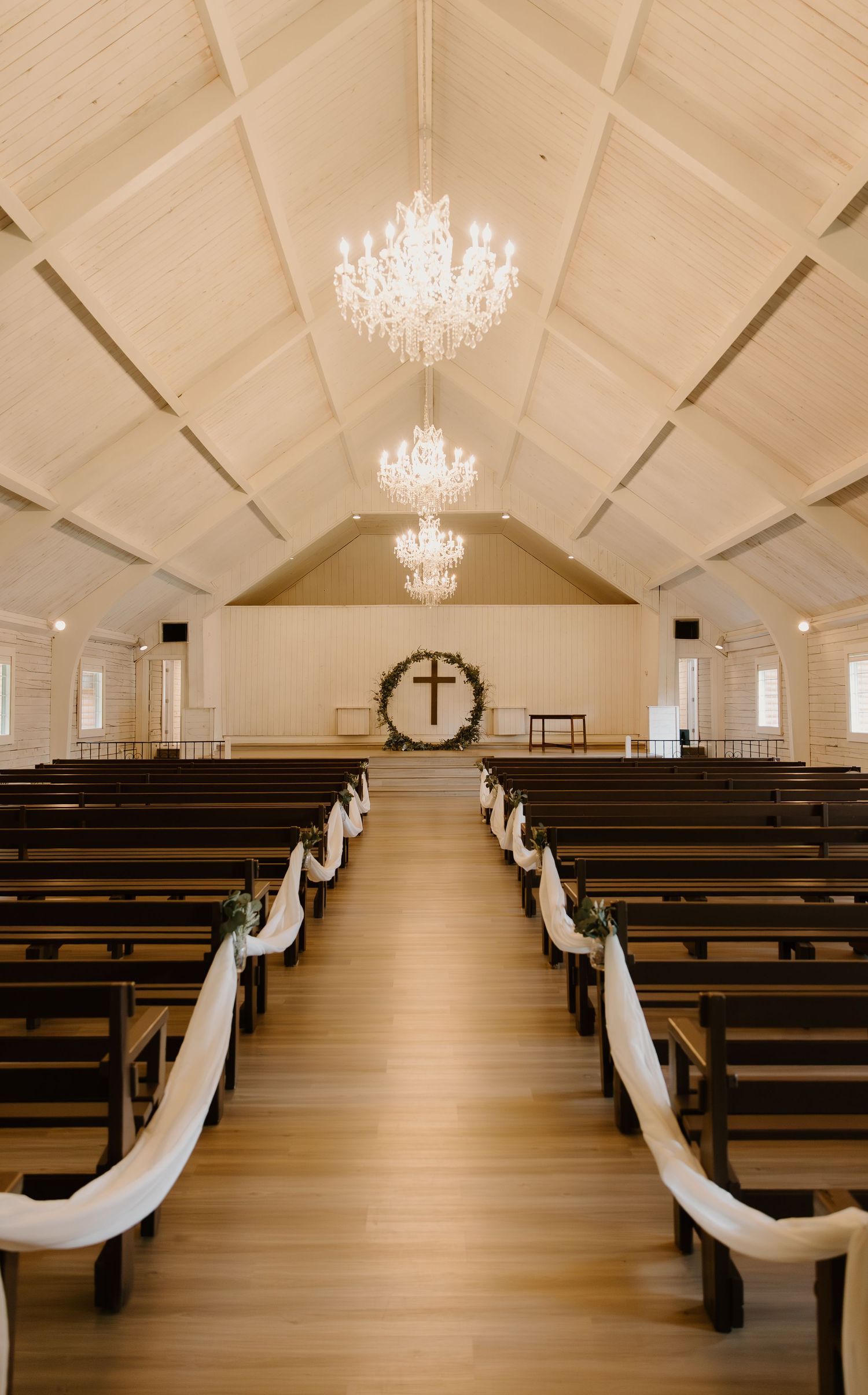 Interior of a white chapel with crystal chandeliers, wooden pews, and white fabric decorations along the center aisle.