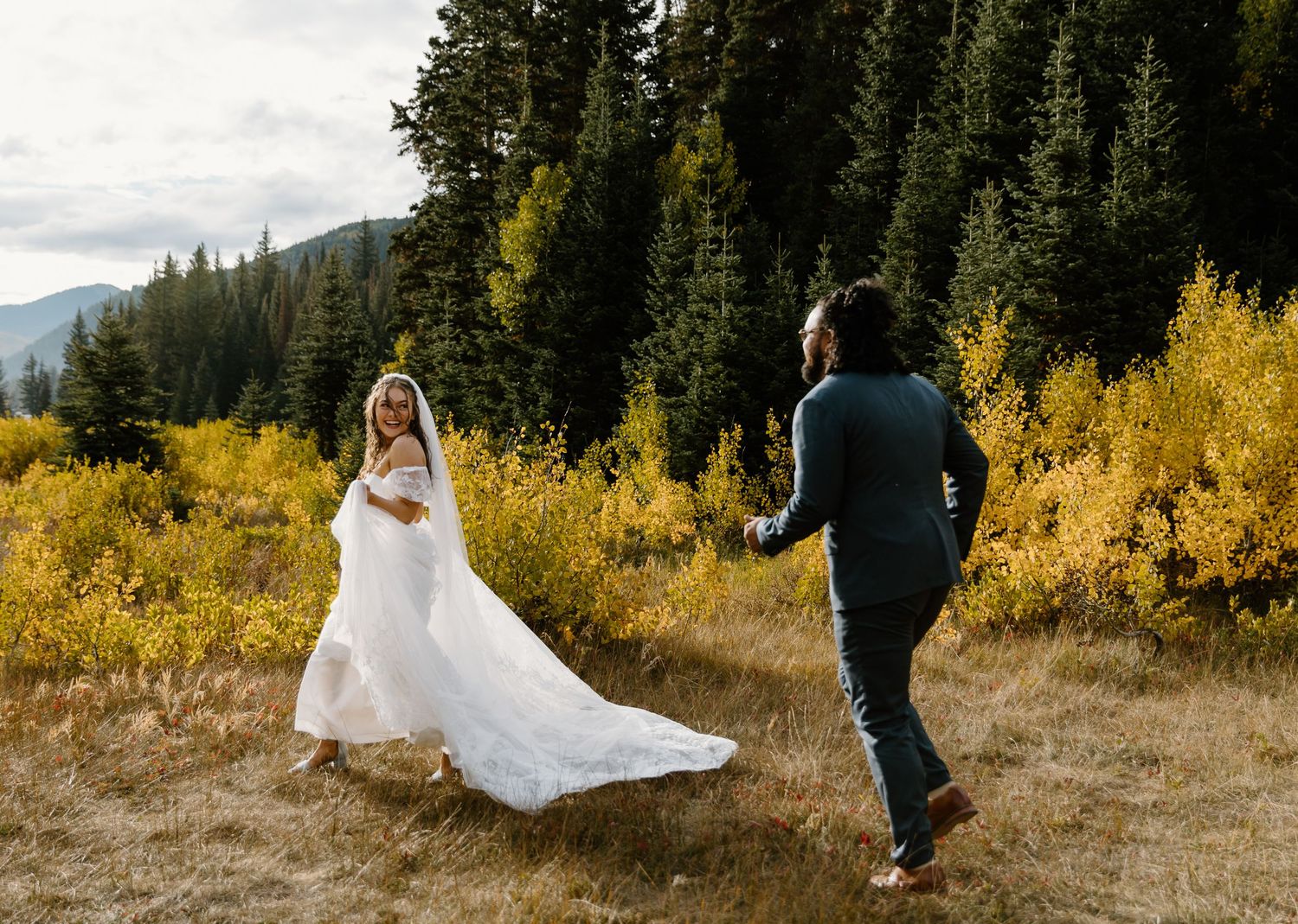 A couple in wedding attire walks through a golden autumn meadow surrounded by evergreen trees in the mountains.