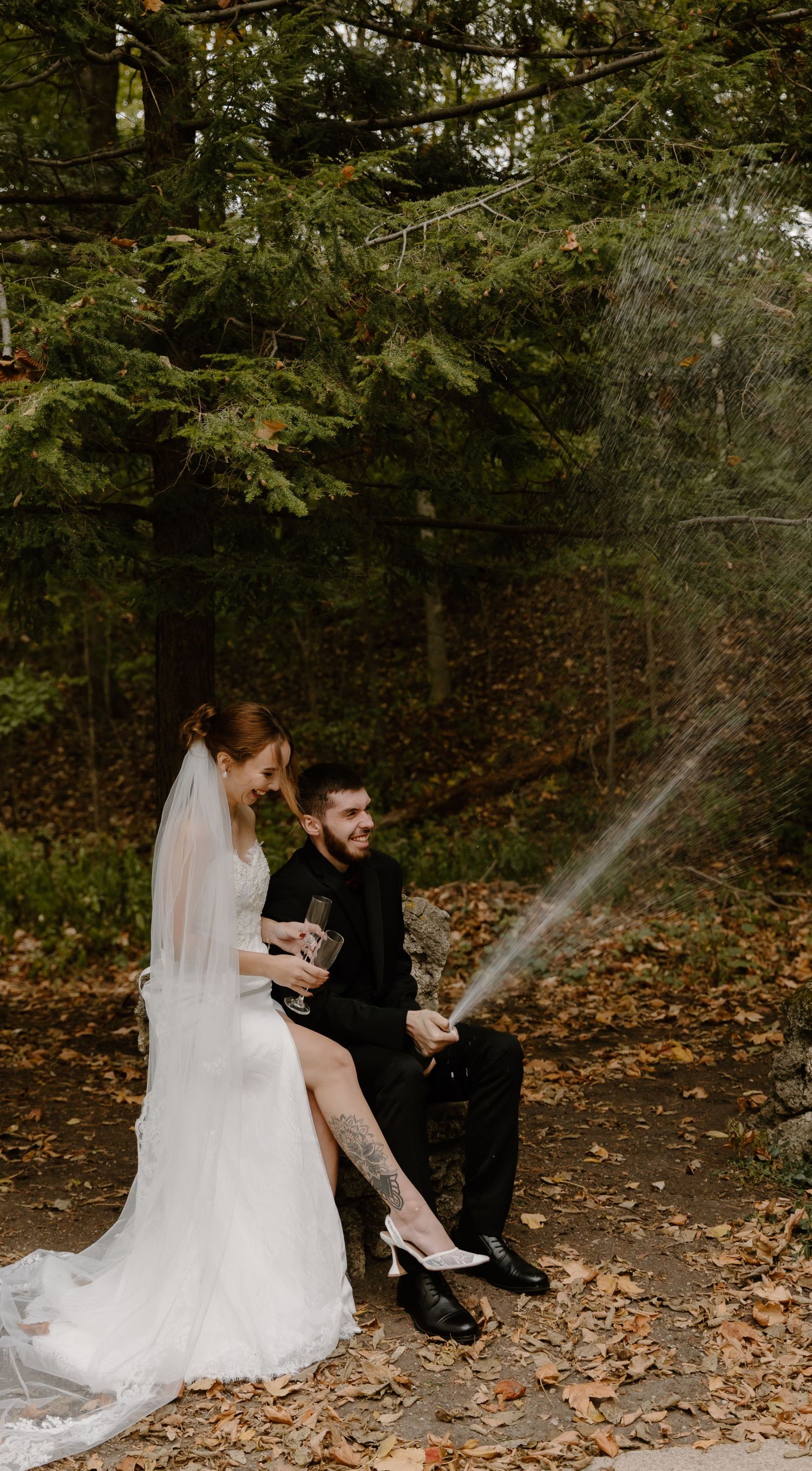 Eloped newlyweds sit together on autumn leaves in a forest setting, sharing champagne.