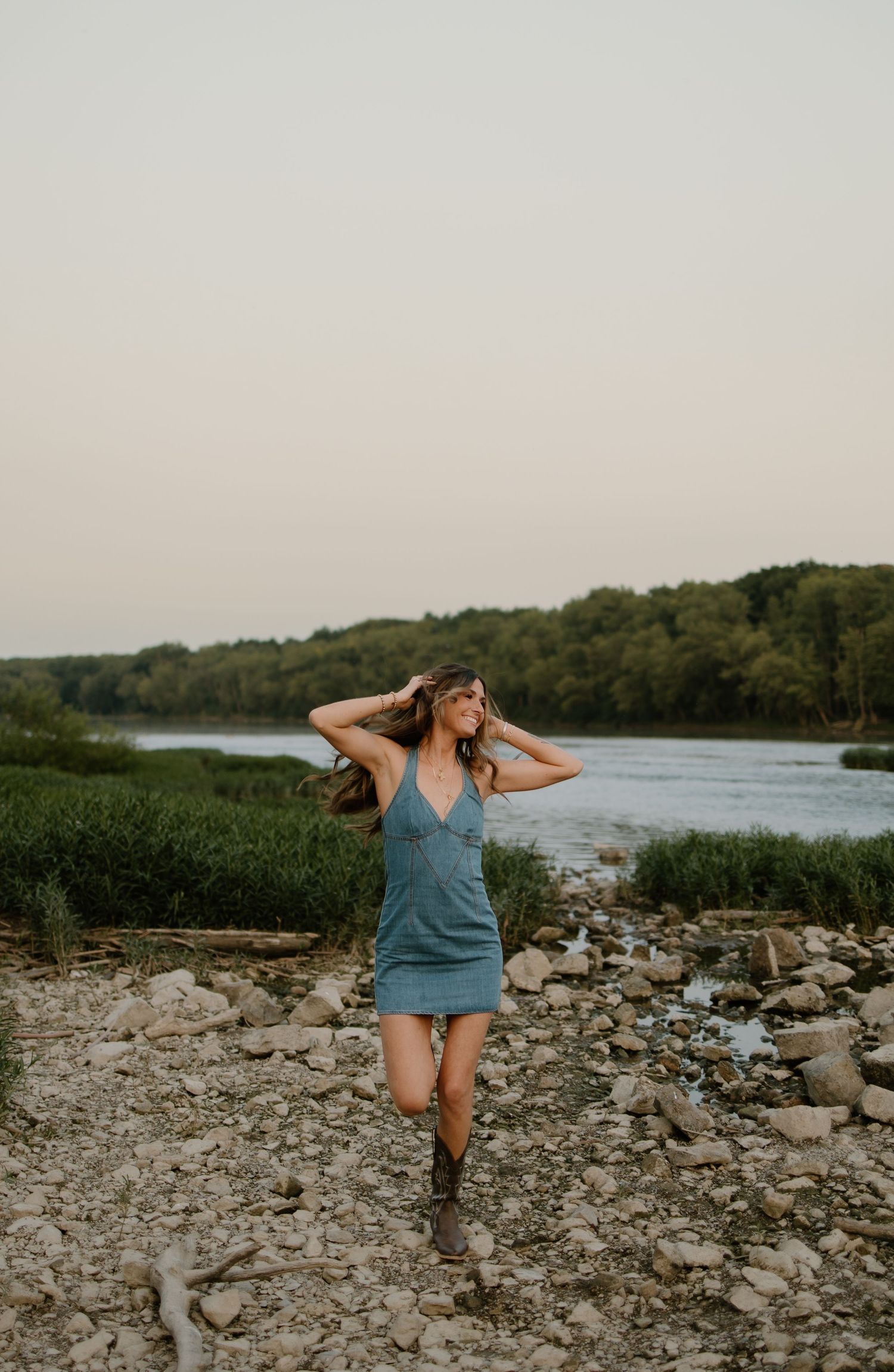 Young woman in blue mini dress and cowboy boots walking along rocky riverbank at sunset.