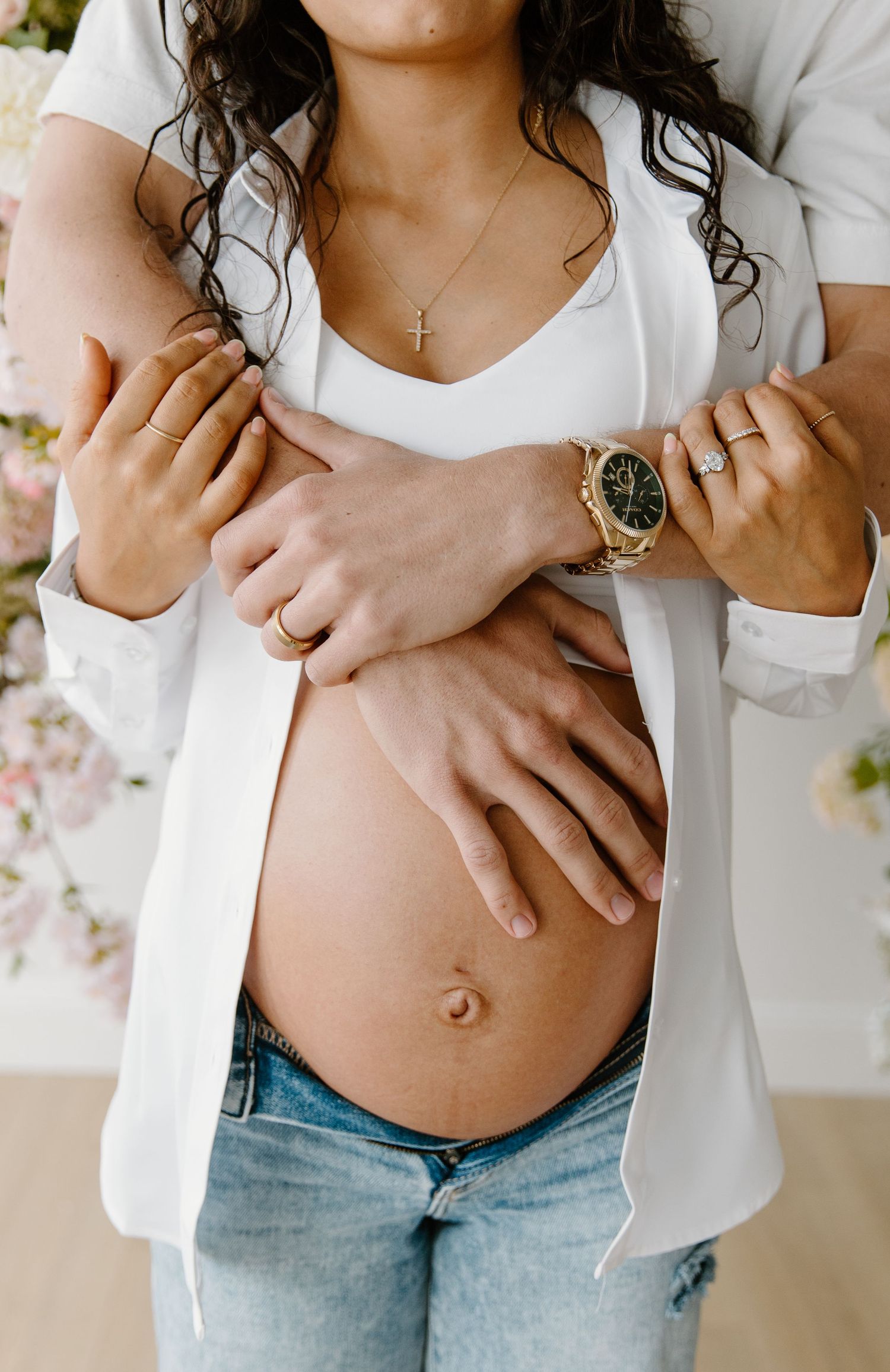 Couple embracing pregnant belly, woman in white blazer and jeans, man's hands with gold watch cradling bump.