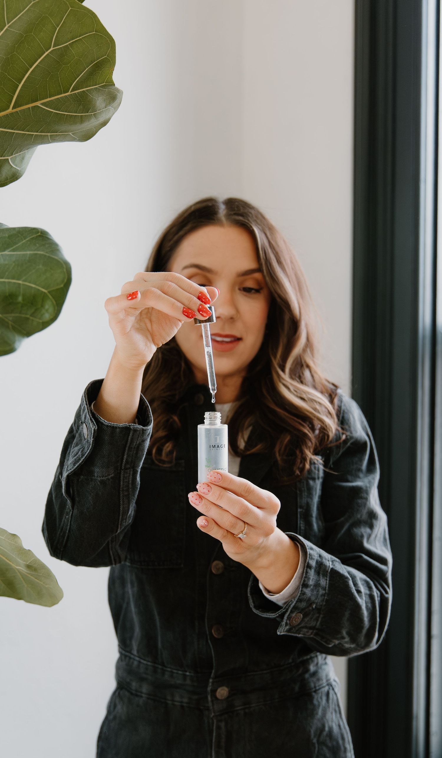 Woman in denim jacket applying facial serum with dropper, red nails, beside green plant.
