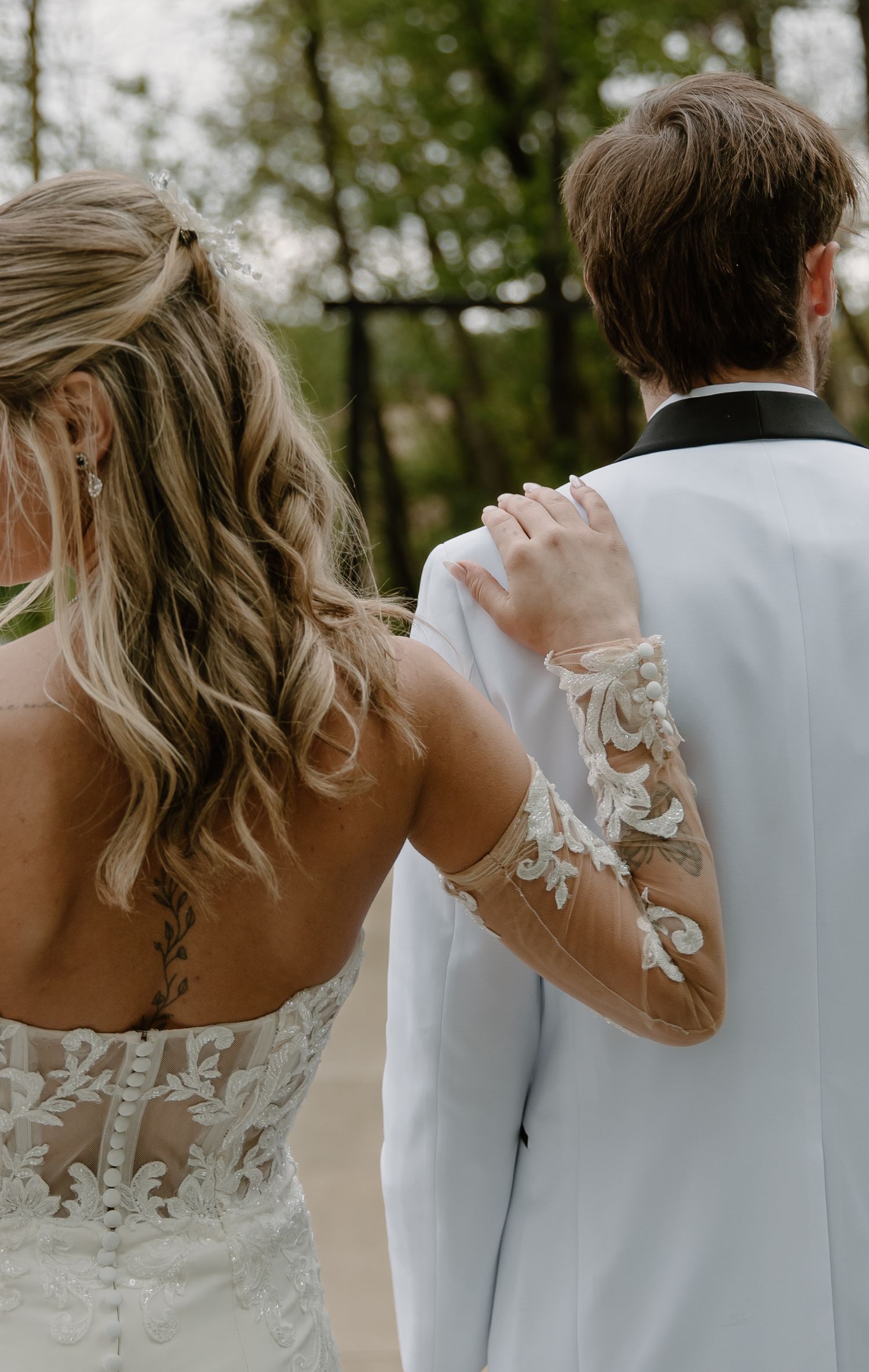Bride in lace wedding gown with long sleeves holds grooms white tuxedo arm from behind outdoors.