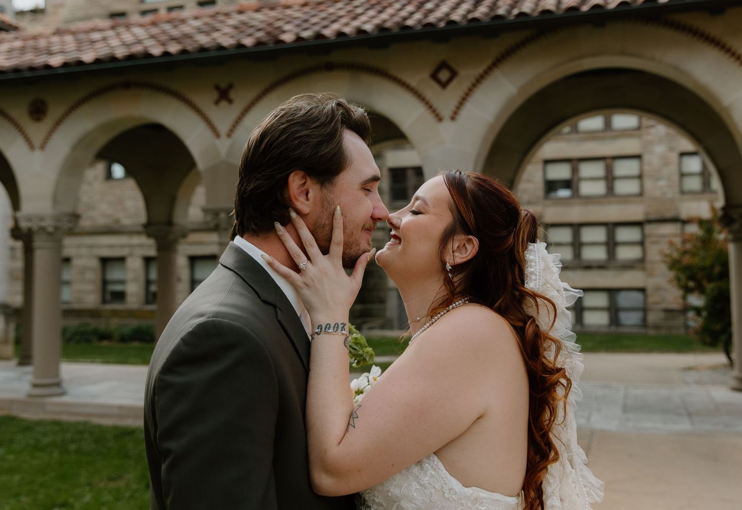 Bride and groom share a romantic almost-kiss moment outdoors near a historic arched stone building on their wedding day.