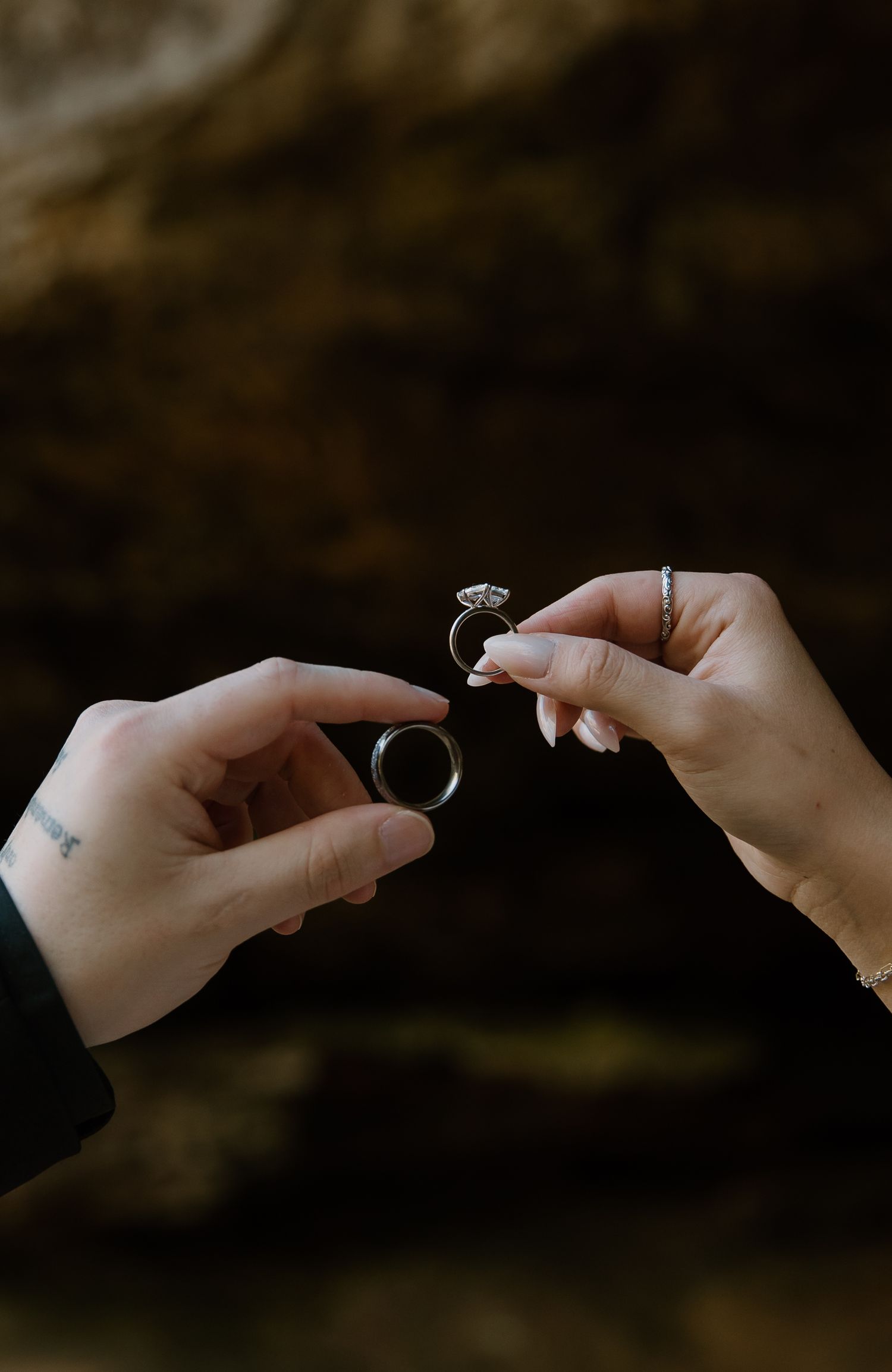Two hands holding wedding rings, one a plain band with a tattoo visible, the other a diamond engagement ring with manicured nails.