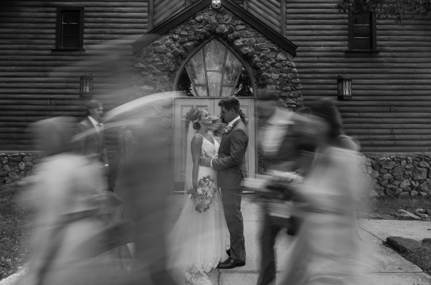 Artistic black and white wedding photo with motion blur of guests walking past couple at church entrance.