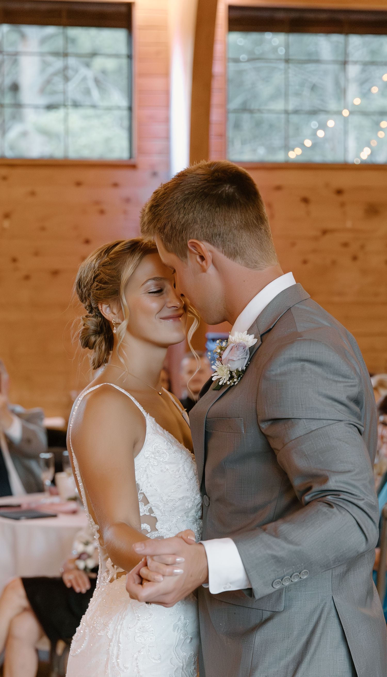 Newlyweds share tender moments during their first dance in rustic wooden venue with twinkling lights.