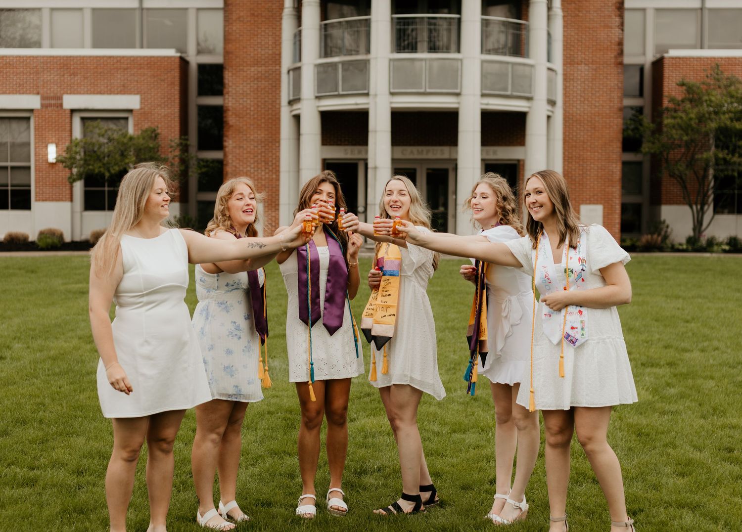 A group of friends in white dresses celebrate together on a grassy lawn in front of a brick building.