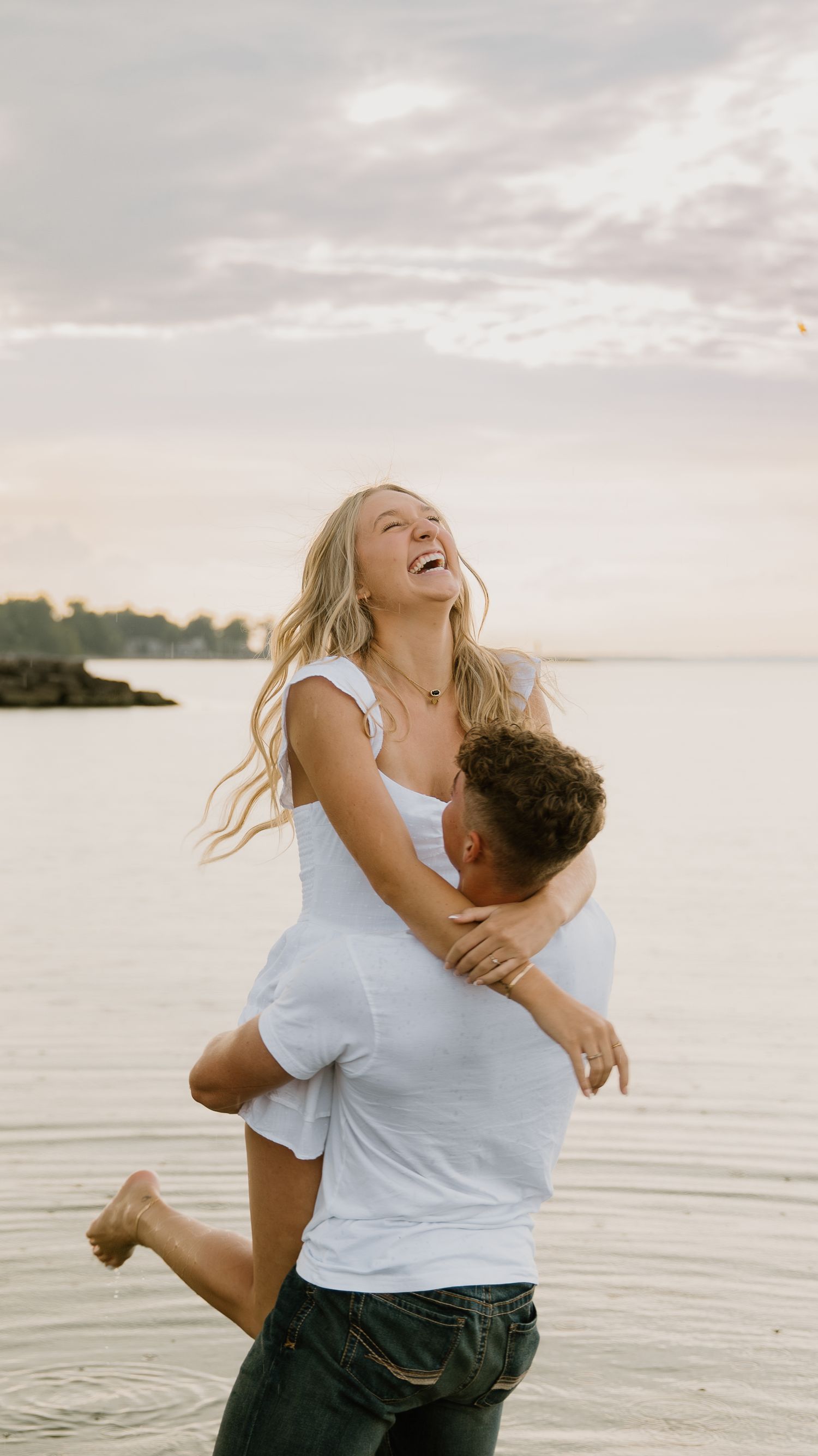 A joyful couple embraces and laughs together by the water at sunset during a romantic photoshoot in Northwest Ohio.