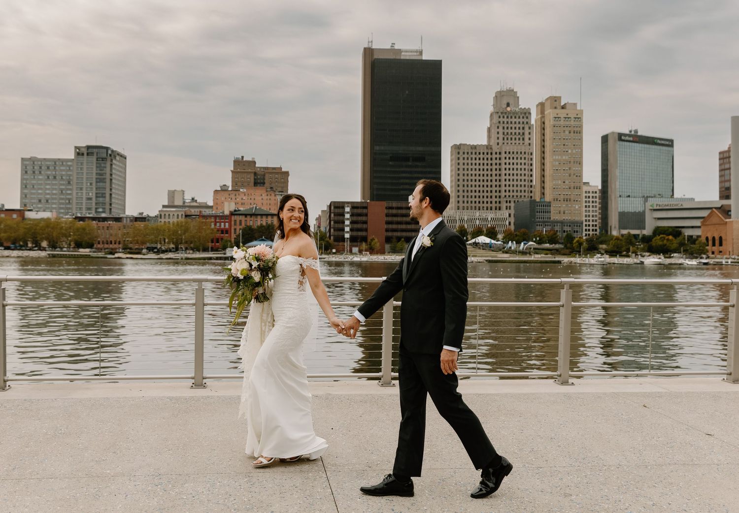 A couple poses for wedding photos on a waterfront with urban buildings and boats visible in the distance.