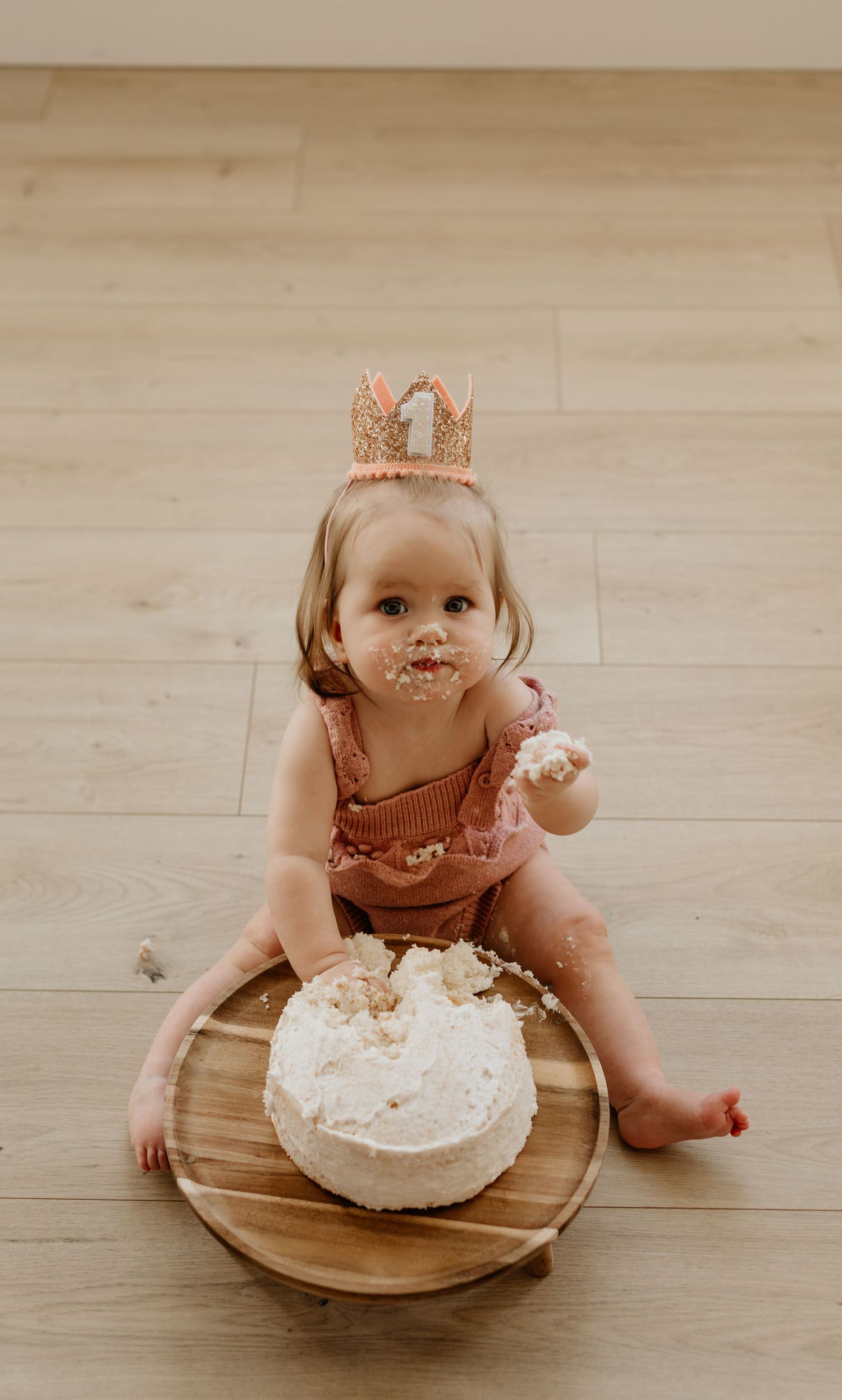 Baby wearing crown sits on wooden floor with white birthday cake.