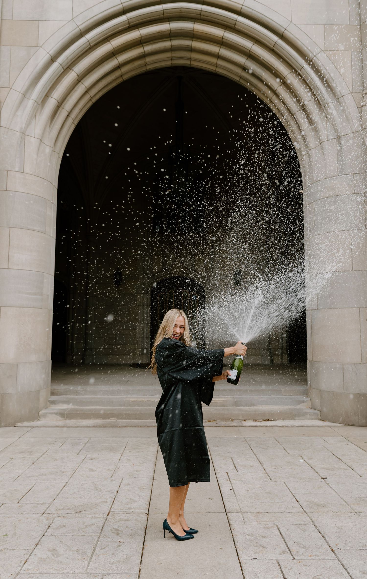 Woman in black coat popping champagne bottle under stone arch, celebrating with bubbly spray outdoors.