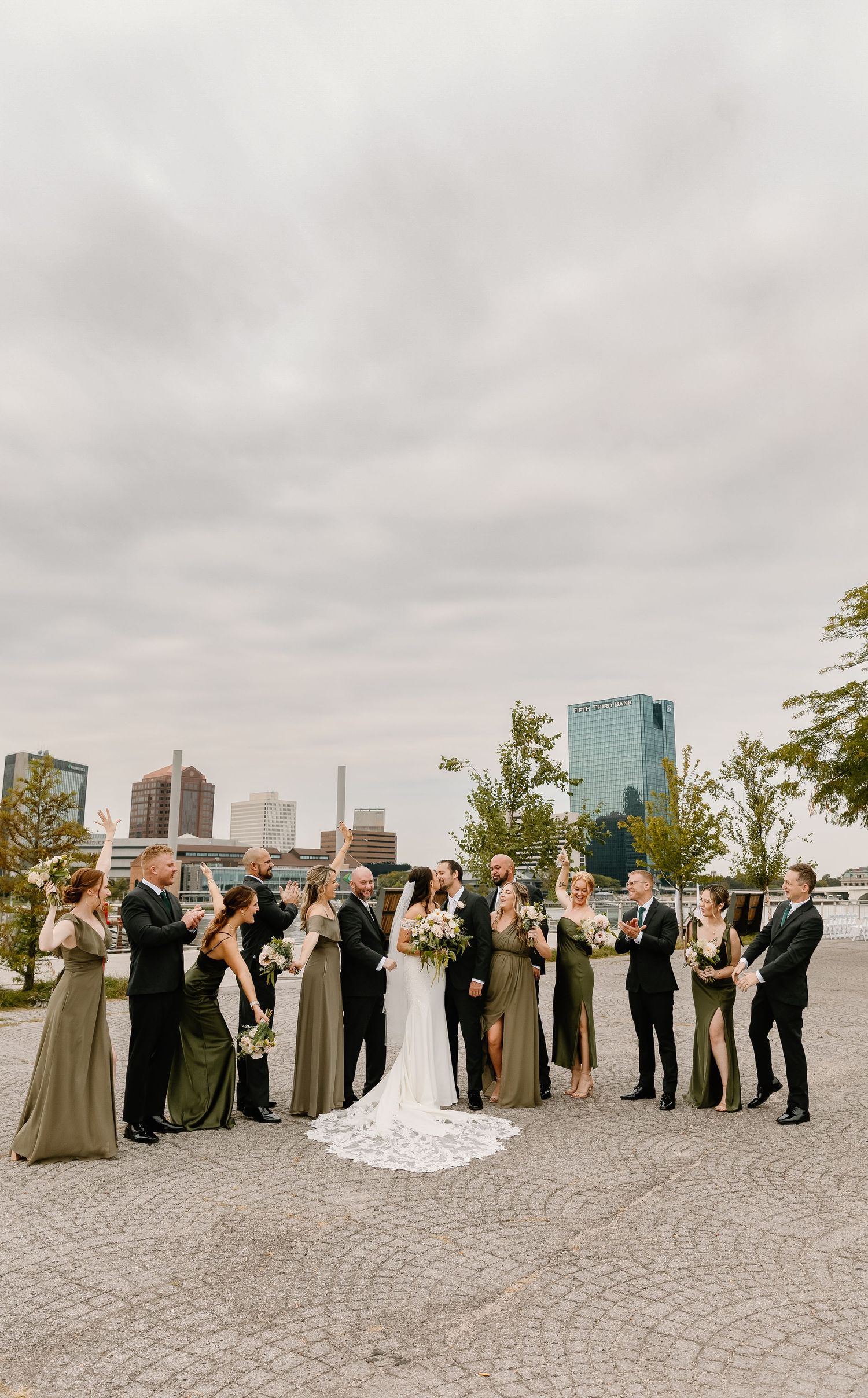 Wedding party lined up for photos on a riverside promenade with city skyline in background.