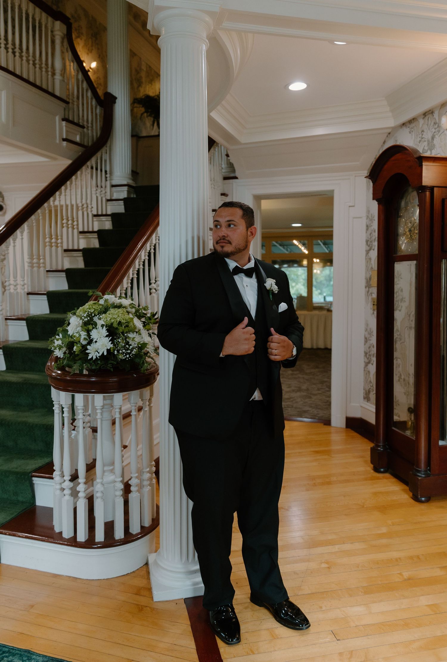 Dapper groom in black tuxedo with boutonniere stands confidently in elegant venue foyer with staircase.